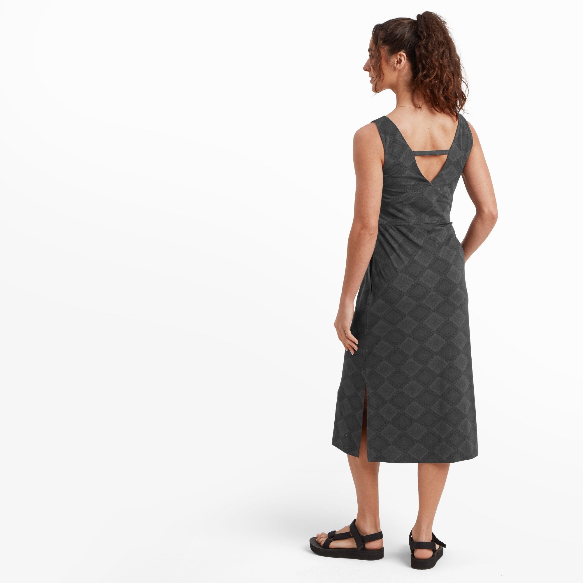 Captures the back design of the Sherpa Adventure Gear Neha Midi Dress in Black, featuring a deep V-back with a thin strap across the shoulders for added structure. The diamond pattern continues seamlessly, highlighting the dress's attention to detail. The model’s hair is styled in a loose ponytail, revealing the back neckline.