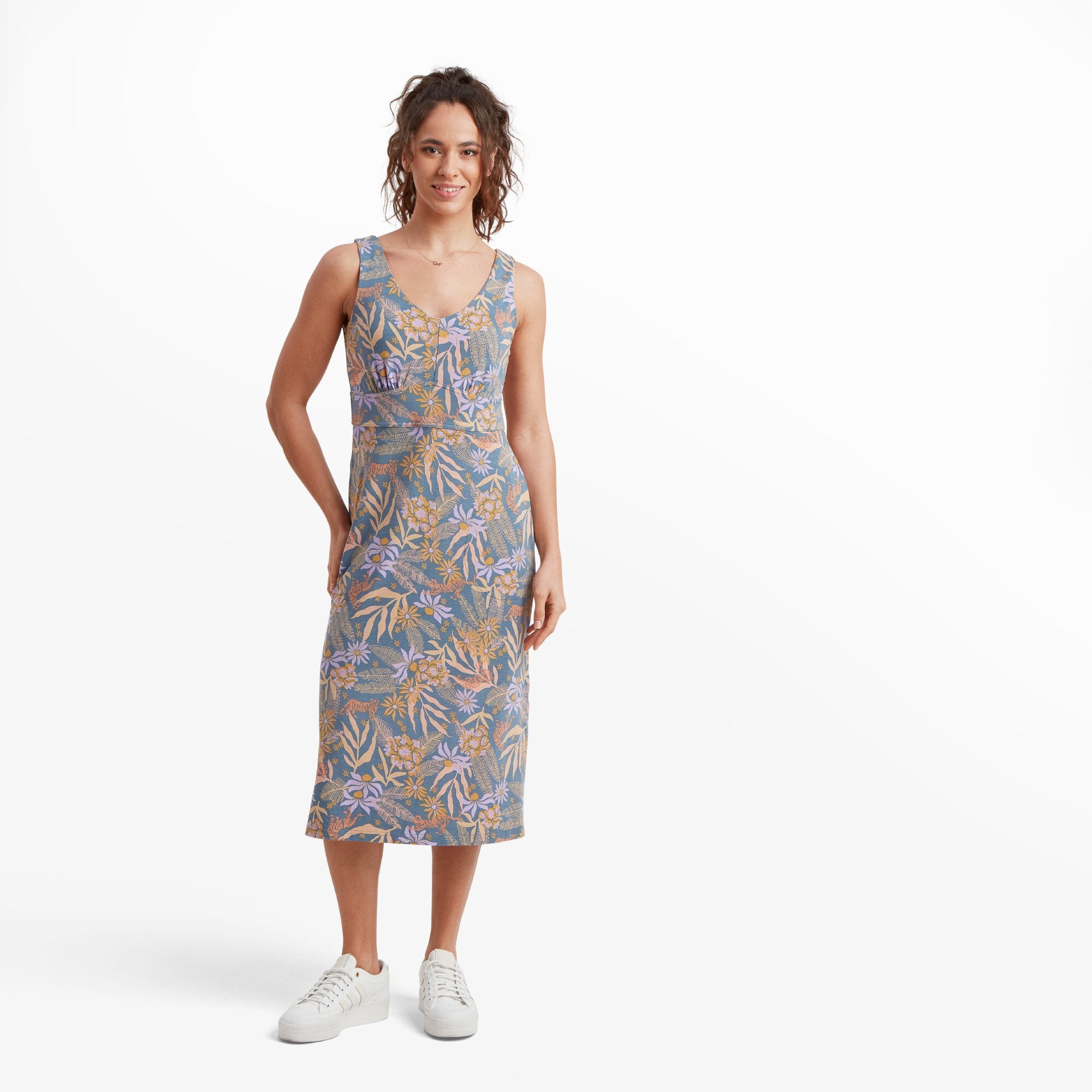 A model is wearing a Sherpa Adventure Gear Neha Midi Dress in Blue in soft pastel tones of blue, pink, and yellow with tiger motifs. The dress has a flattering V-neckline, a fitted waist, and a slightly flared hem, paired with white trainers for a casual look.