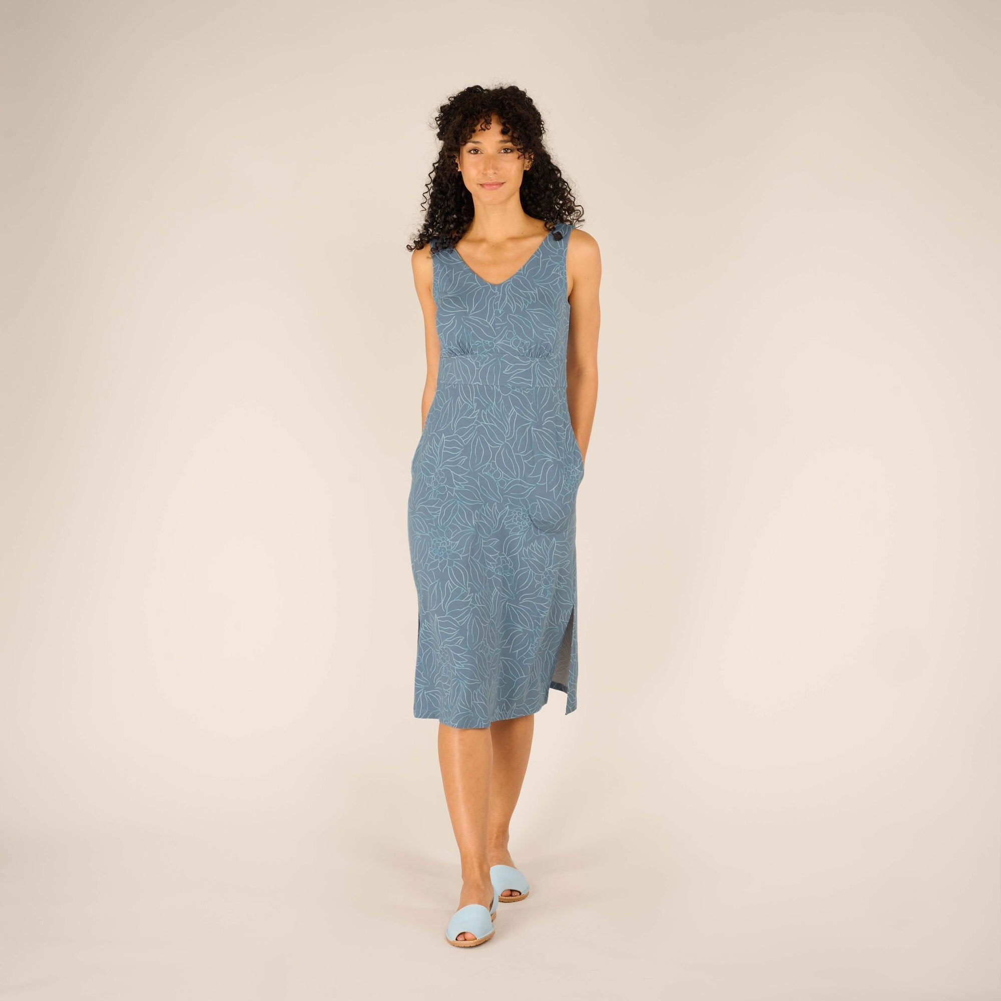 A woman with curly black hair and a warm smile is standing against a neutral beige background, wearing a Sherpa Adventure Gear Neha Midi Dress in Blue with a subtle floral pattern. The dress has a V-neckline and thick shoulder straps, and it flows down to her mid-calf with a slight slit on one side. She is wearing light blue open-toe sandals and has her hands tucked into the dress’s side pockets.