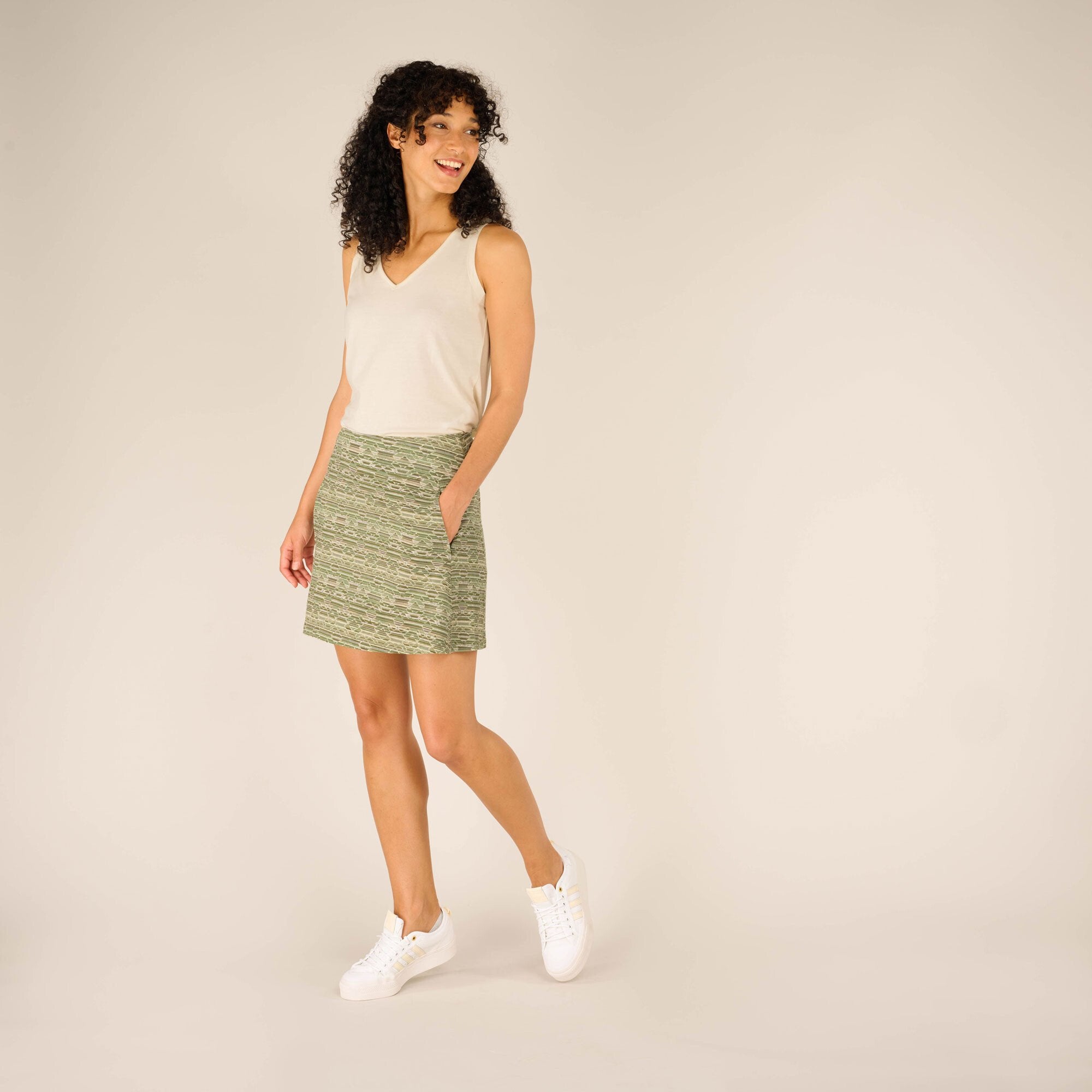 A full-body shot of a woman with curly hair modeling the Sherpa Adventure Gear Neha Skort in Green. She pairs it with a white sleeveless top and white trainers, standing with one foot slightly lifted, smiling.