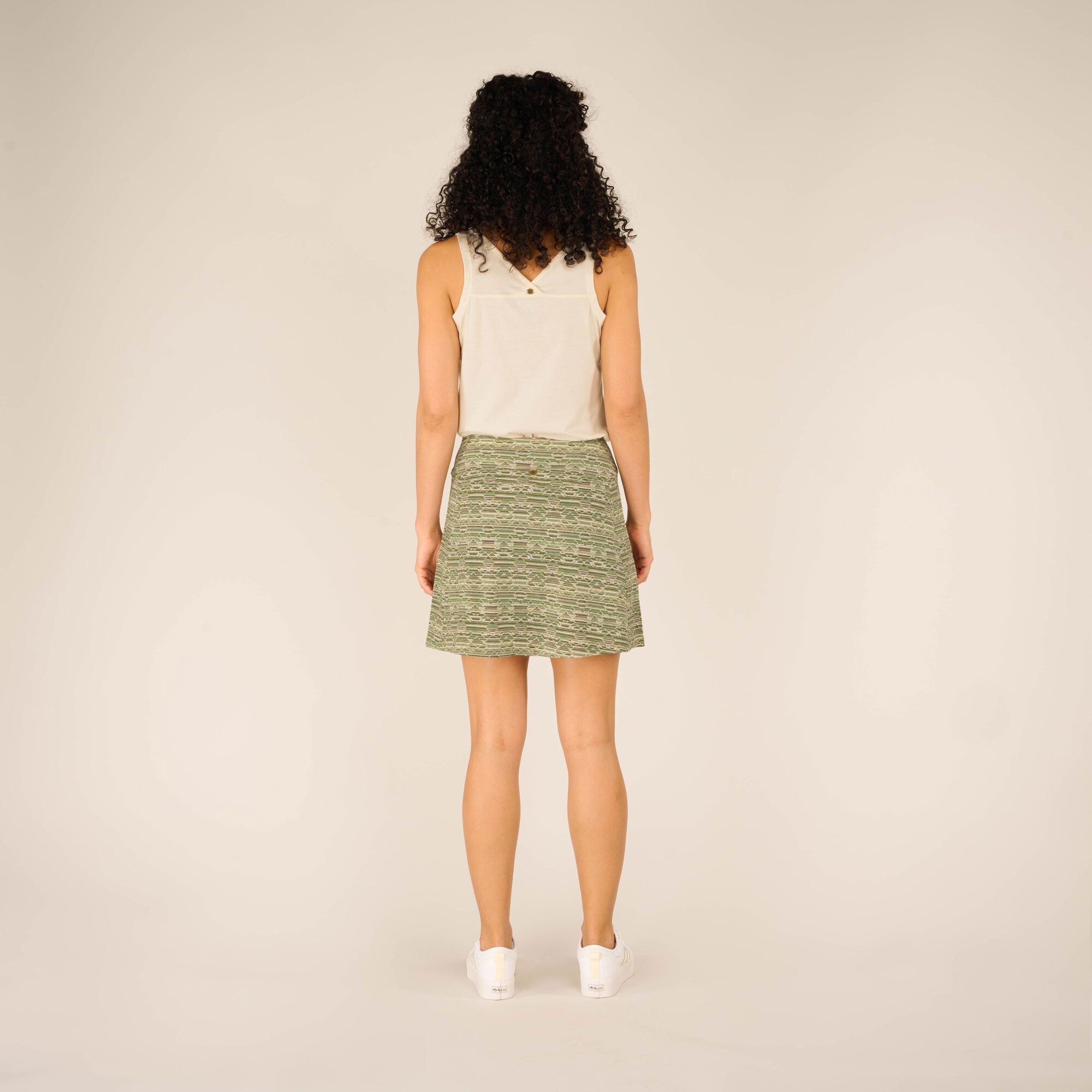 A back view of the Sherpa Adventure Gear Neha Skort in Green, showing its length and fit. The model's white sleeveless top has a small keyhole detail at the upper back. She stands with her hands at her sides.