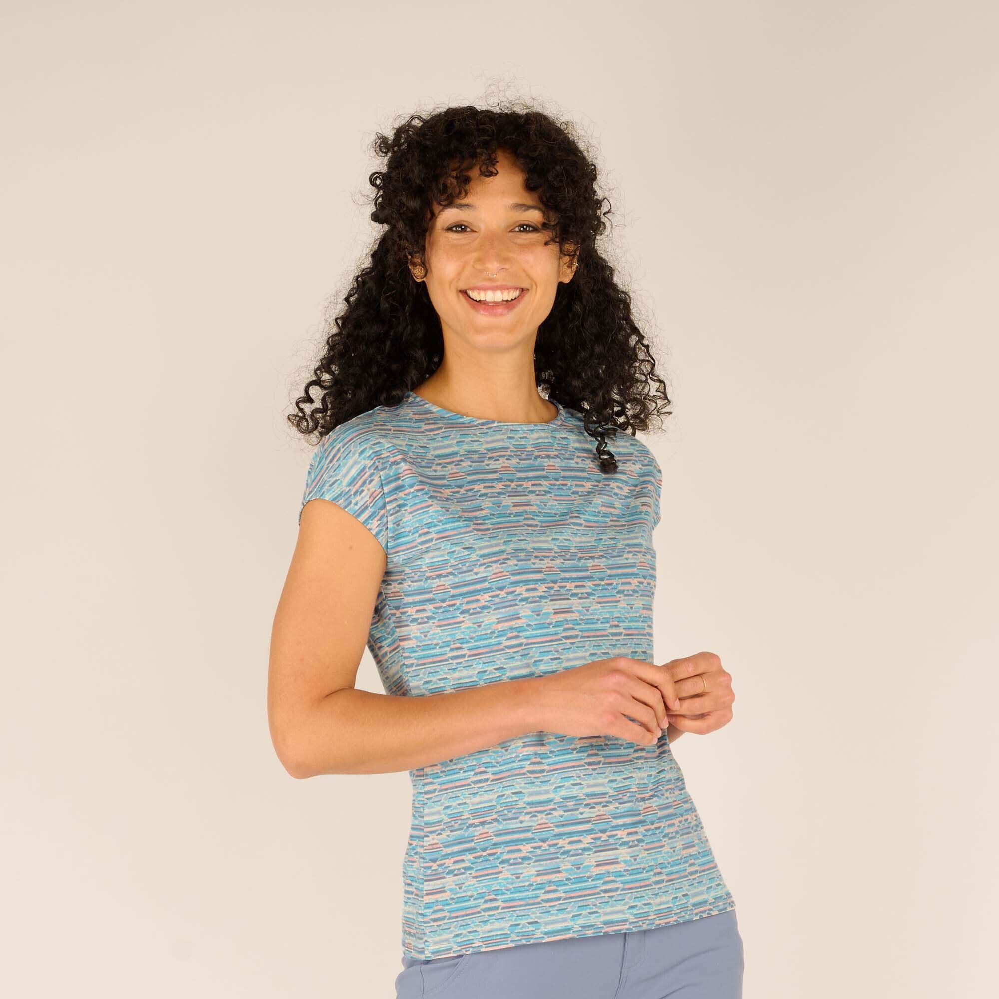 A woman with curly black hair smiles brightly while wearing a Sherpa Adventure Gear Neha T-Shirt in Blue. The lightweight fabric has an abstract wave-like design in shades of blue, white, and pink. She stands against a neutral beige background, her posture relaxed with hands gently clasped.