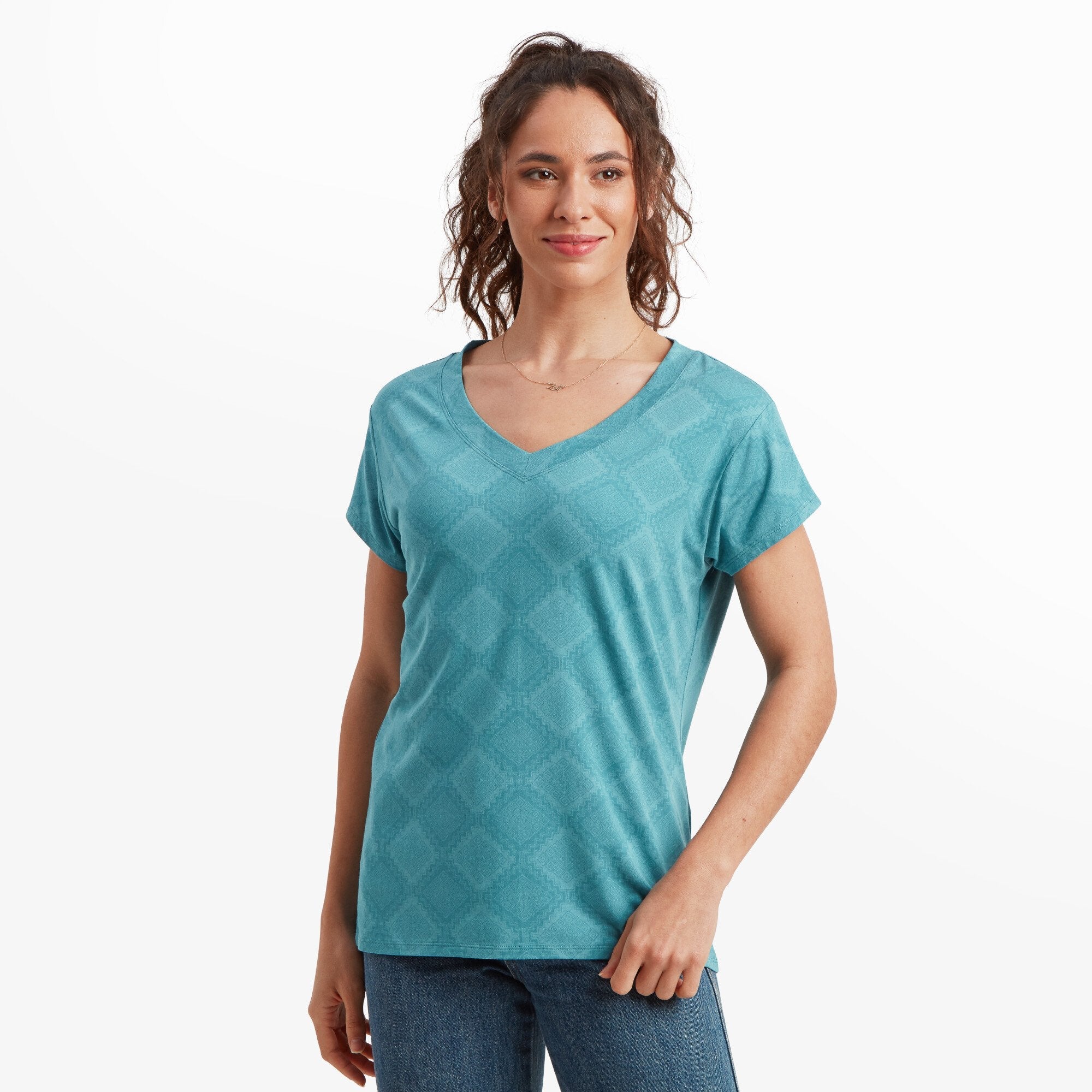 A woman with shoulder-length curly brown hair, light tan skin, and a smile is wearing a Sherpa Adventure Gear Neha V-Neck T-Shirt in Blue with a subtle geometric pattern and short sleeves. She is standing in a relaxed pose with her hands at her sides, looking slightly off to the side. She is also wearing blue jeans. The background is a plain white setting.