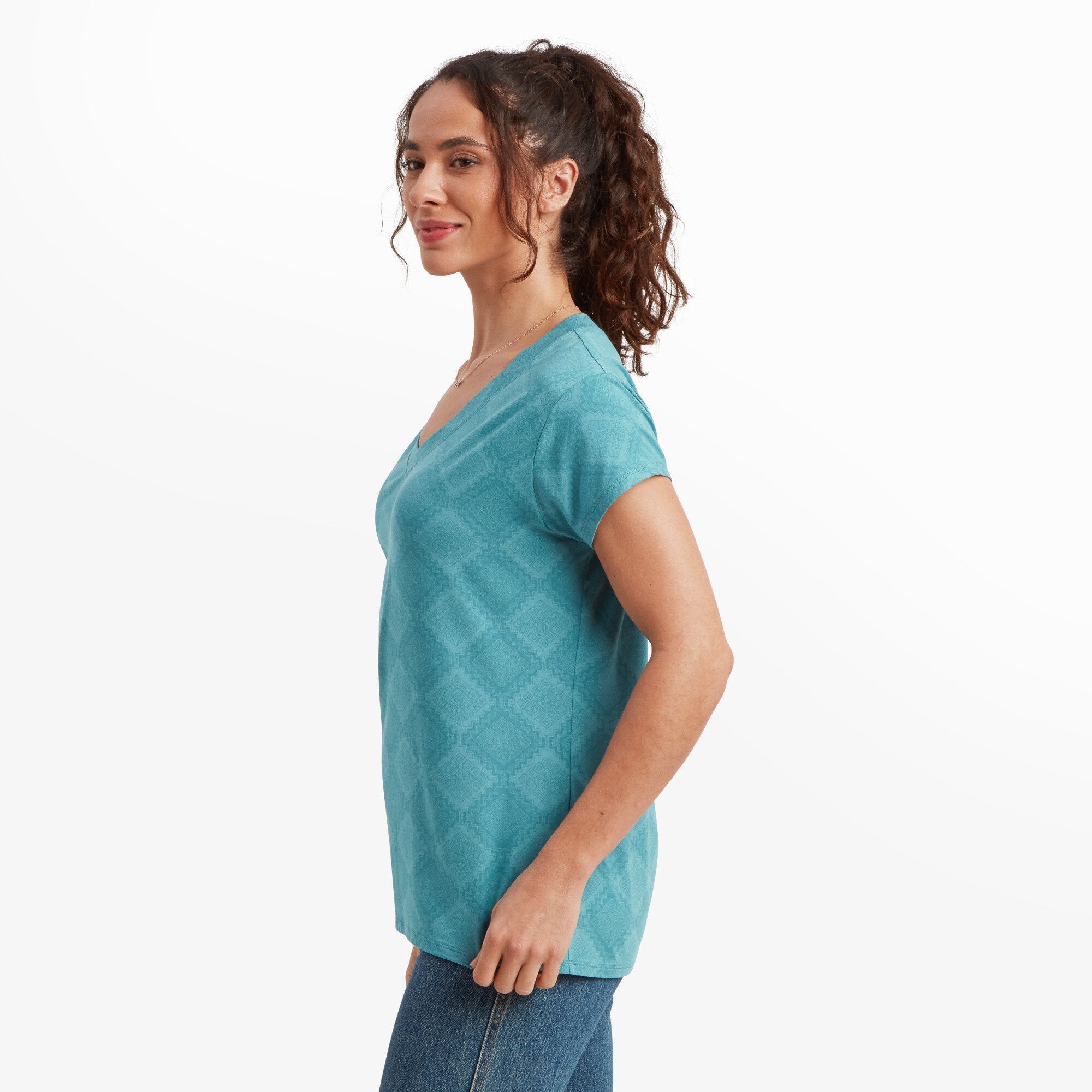 A side profile of the same woman wearing the Sherpa Adventure Gear Neha V-Neck T-Shirt in Blue and jeans. She is smiling gently while looking slightly toward the camera. The geometric pattern on the fabric is faintly visible. Her curly brown hair is tied back into a high ponytail.