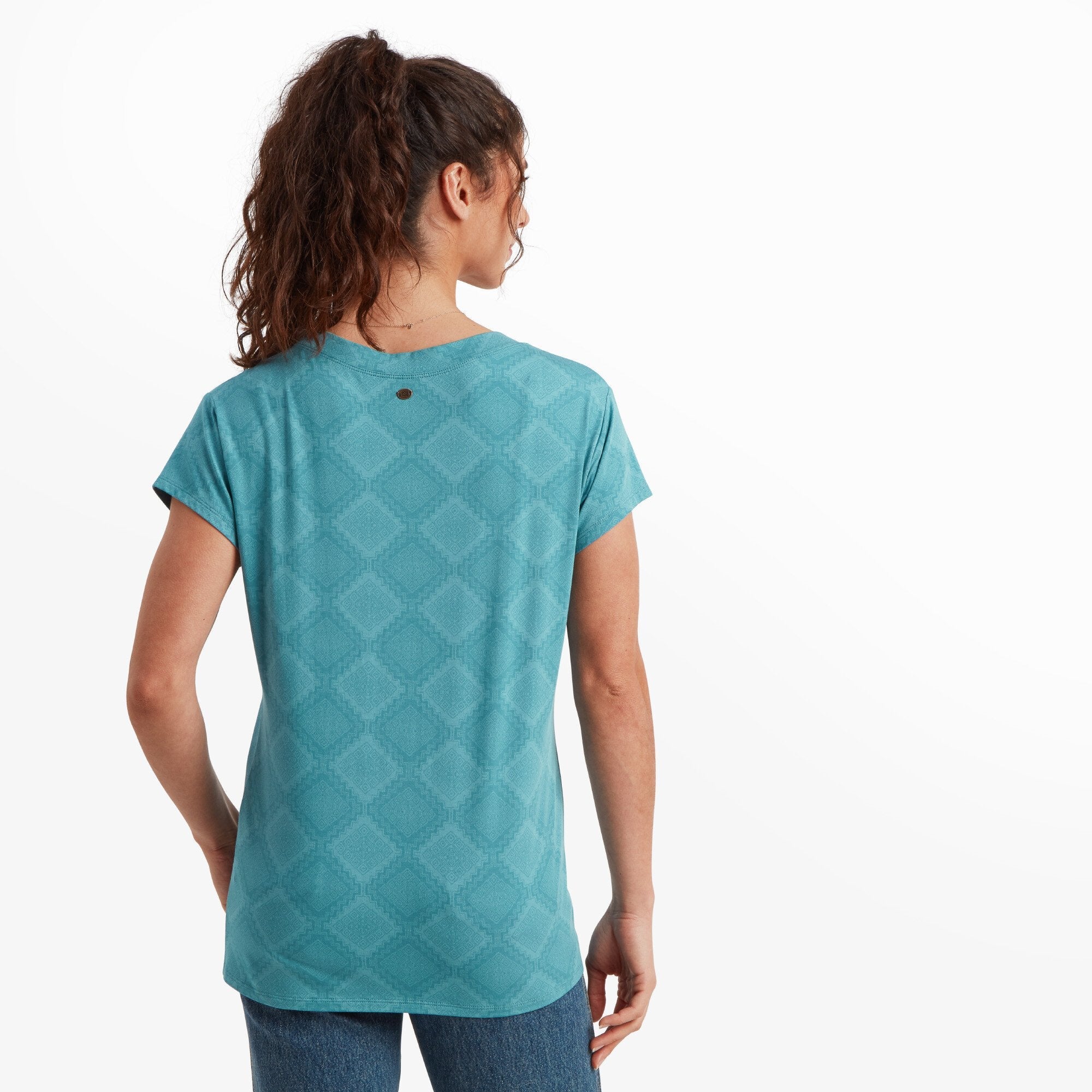 A back view of the woman wearing the Sherpa Adventure Gear Neha V-Neck T-Shirt in Blue. She is standing with her hands at her sides, showing the back details of the shirt, which include a subtle geometric pattern and a small decorative button near the upper center back. Her curly hair is tied back, revealing the back neckline of the shirt.