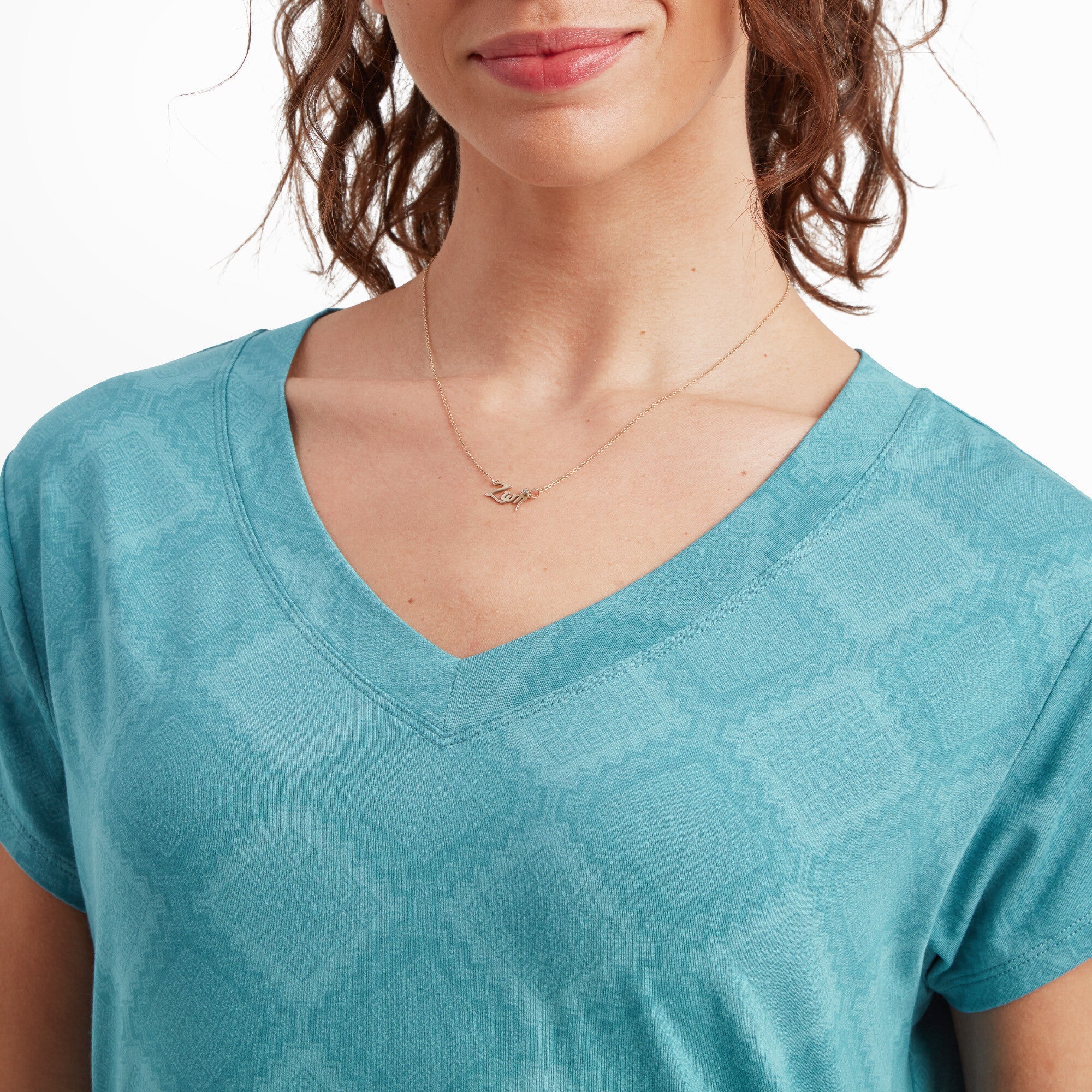 A close-up of the woman’s upper chest and neck, highlighting the V-neckline of the Sherpa Adventure Gear Neha V-Neck T-Shirt in Blue. The subtle geometric pattern is more visible in this view. She is wearing a delicate gold necklace with a small script pendant that says "love." The texture and stitching of the fabric are clearly visible in this close-up.