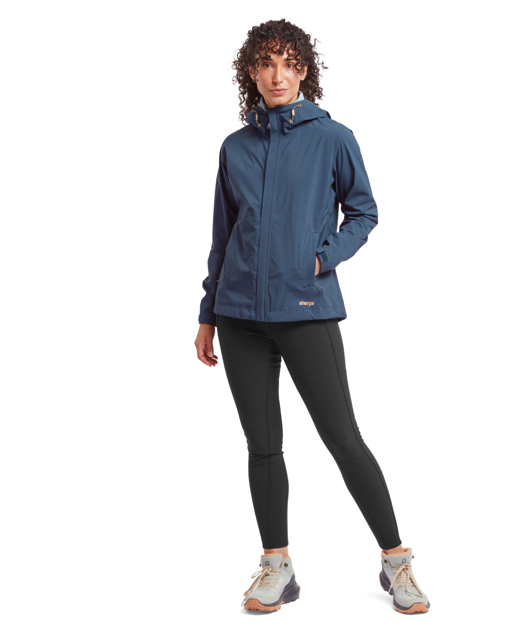 A full-body shot of the model wearing the Sherpa Adventure Gear Nima 2.5-Layer Jacket in Blue paired with black leggings and trail trainers. The model stands in a slight pose, showing the jacket's slim fit and versatility for outdoor activities.