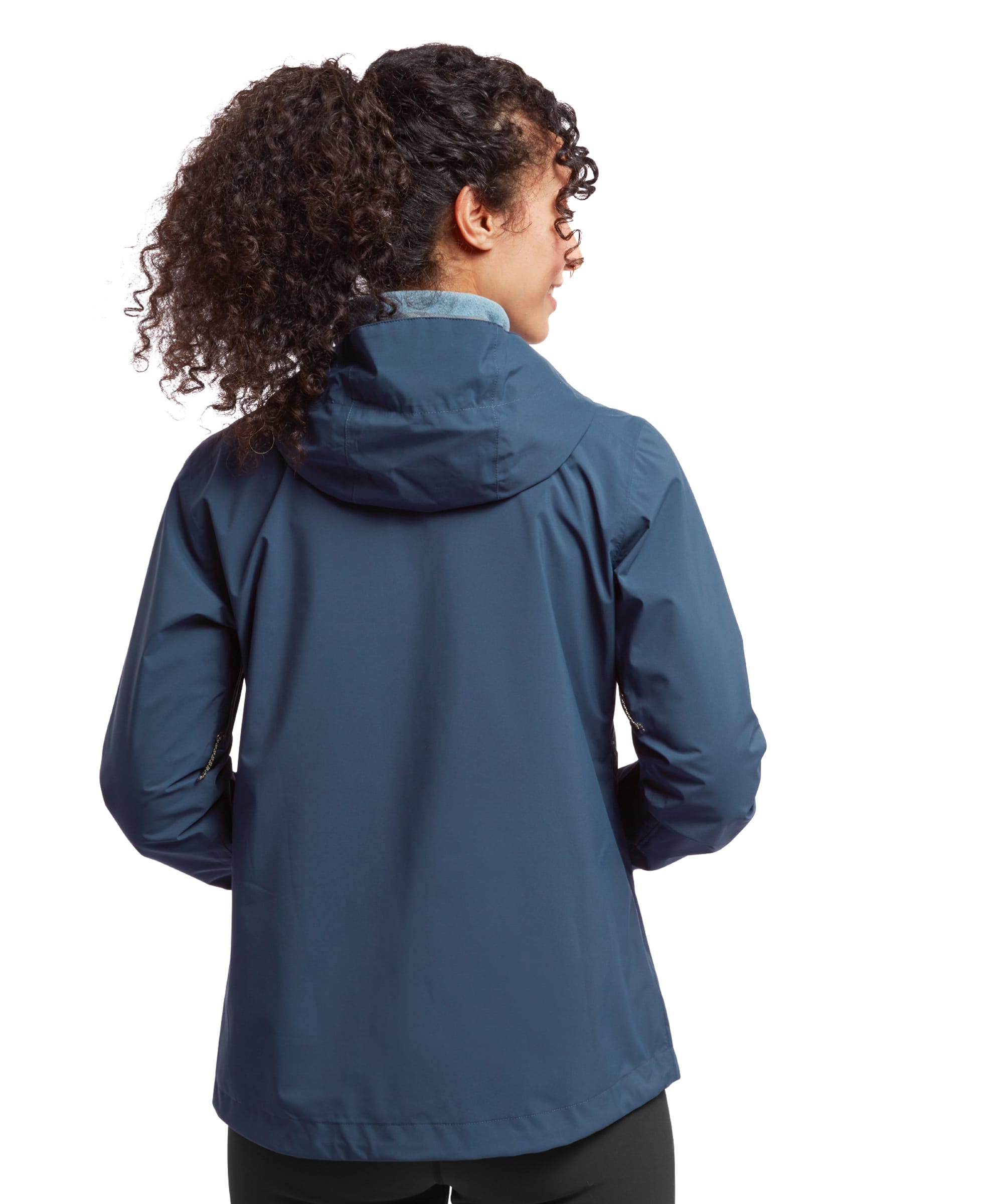 A rear view of the Sherpa Adventure Gear Nima 2.5-Layer Jacket in Blue, displaying its seamless design and hood lying flat on the model's back. The model's hands are casually placed behind her, with her curly ponytail adding contrast to the clean lines of the jacket.