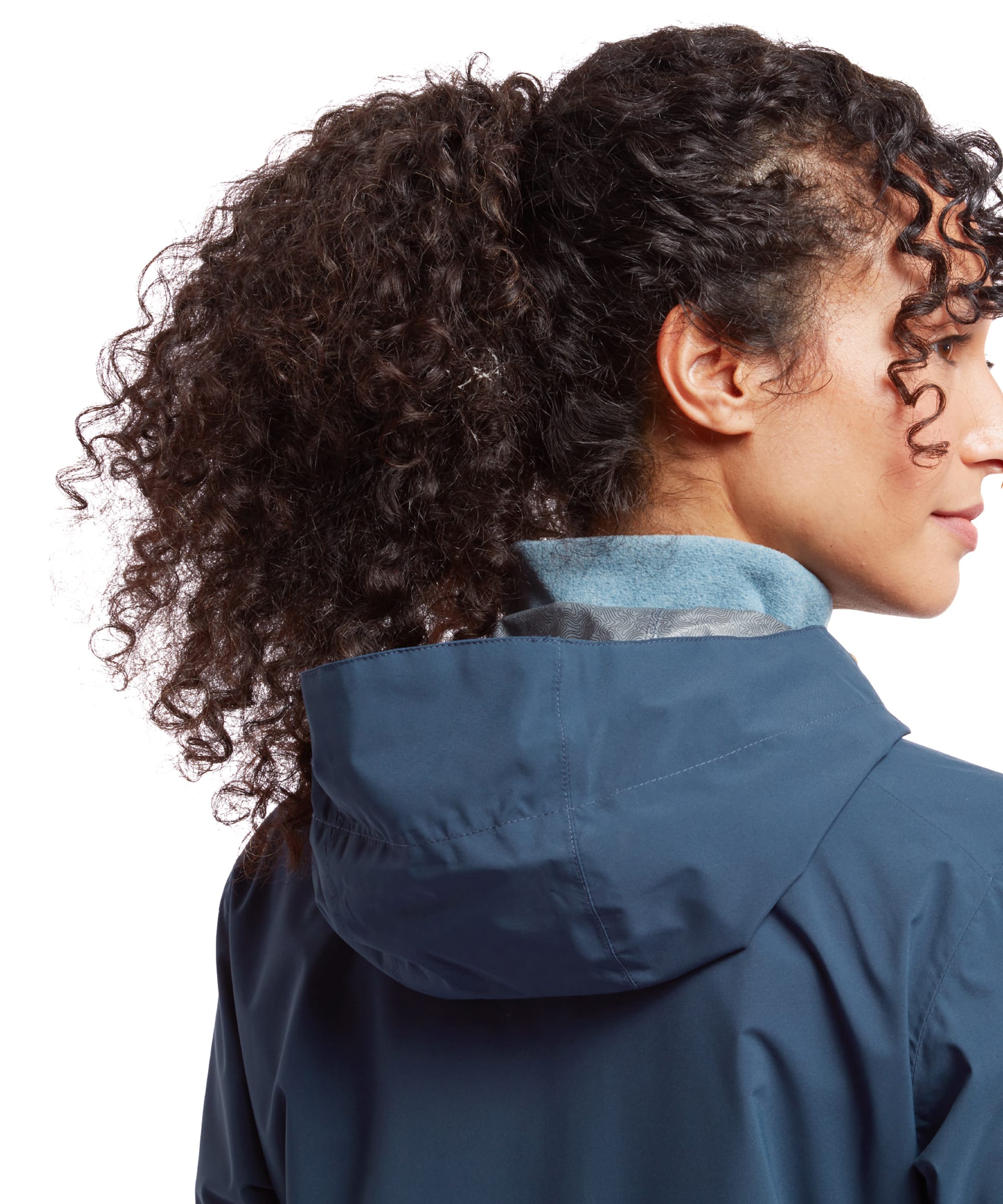 The model is shown from the side with a focus on the hood and upper back of the Sherpa Adventure Gear Nima 2.5-Layer Jacket in Blue. The model's curly ponytail cascades over the hood, which frames the neckline comfortably with a glimpse of a soft fleece lining.