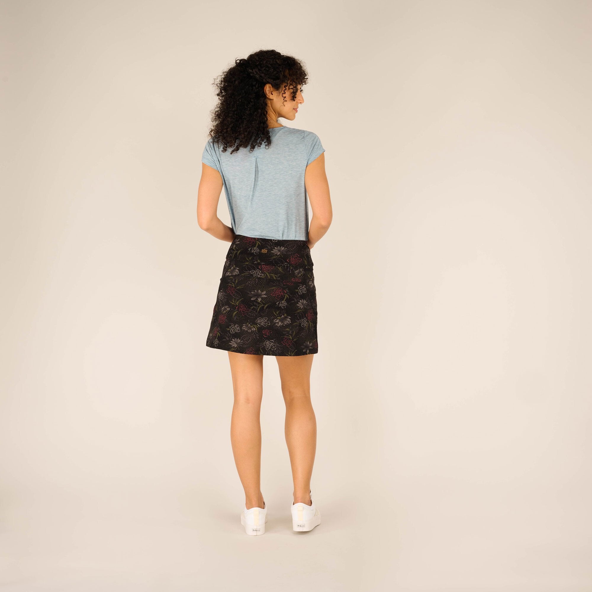 A rear view of the same outfit, showcasing the back design of the light blue t-shirt, which falls slightly loose over the floral-patterned Sherpa Adventure Gear Nisha Skort in Black. The skort fits snugly at the waist and flares slightly towards the hem. Her hair is left loose, and she pairs the outfit with white trainers, giving a casual and comfortable vibe. The neutral background emphasises the outfit's design and details.