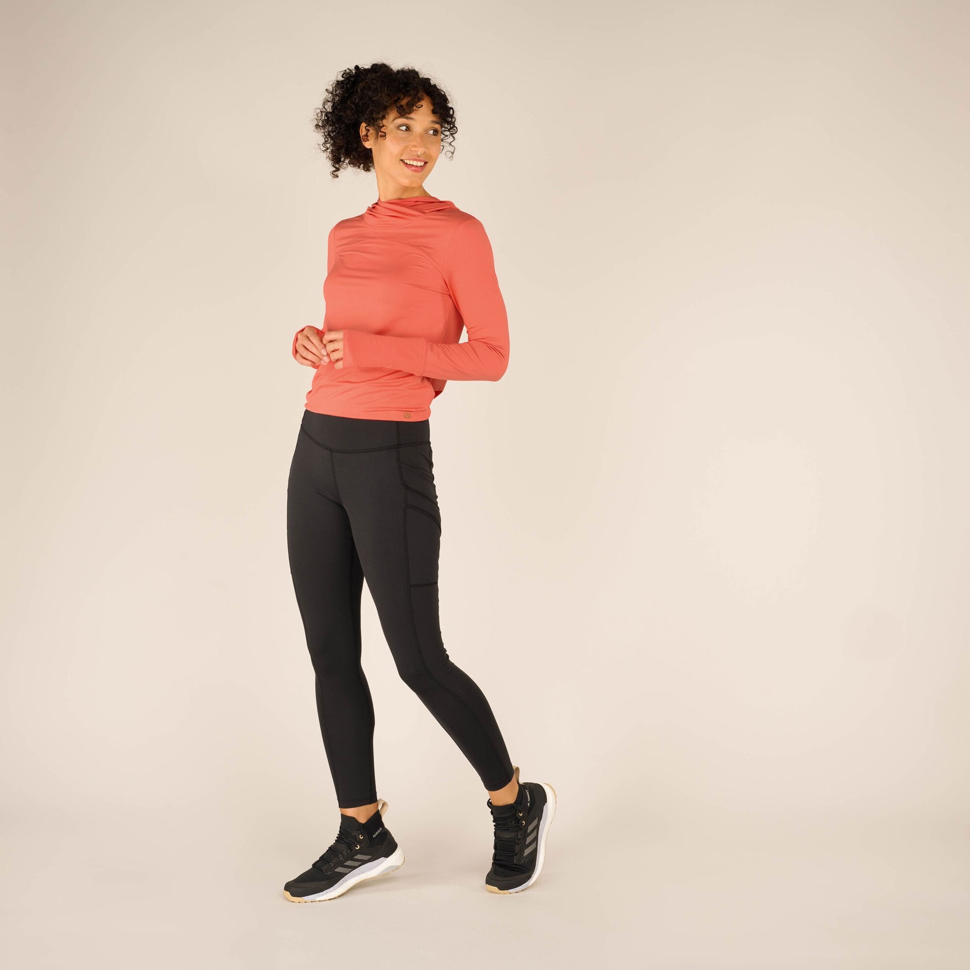 Full-body view of a model in a coral long-sleeve athletic top and Sherpa Adventure Gear Nisha Tight in Black. The model stands at a slight angle with one foot slightly forward and hands relaxed at the sides. The black leggings are high-waisted and curve along the legs, appearing sleek and functional. The bright coral top contrasts with the black leggings. The model smiles confidently while wearing black athletic shoes with white soles.