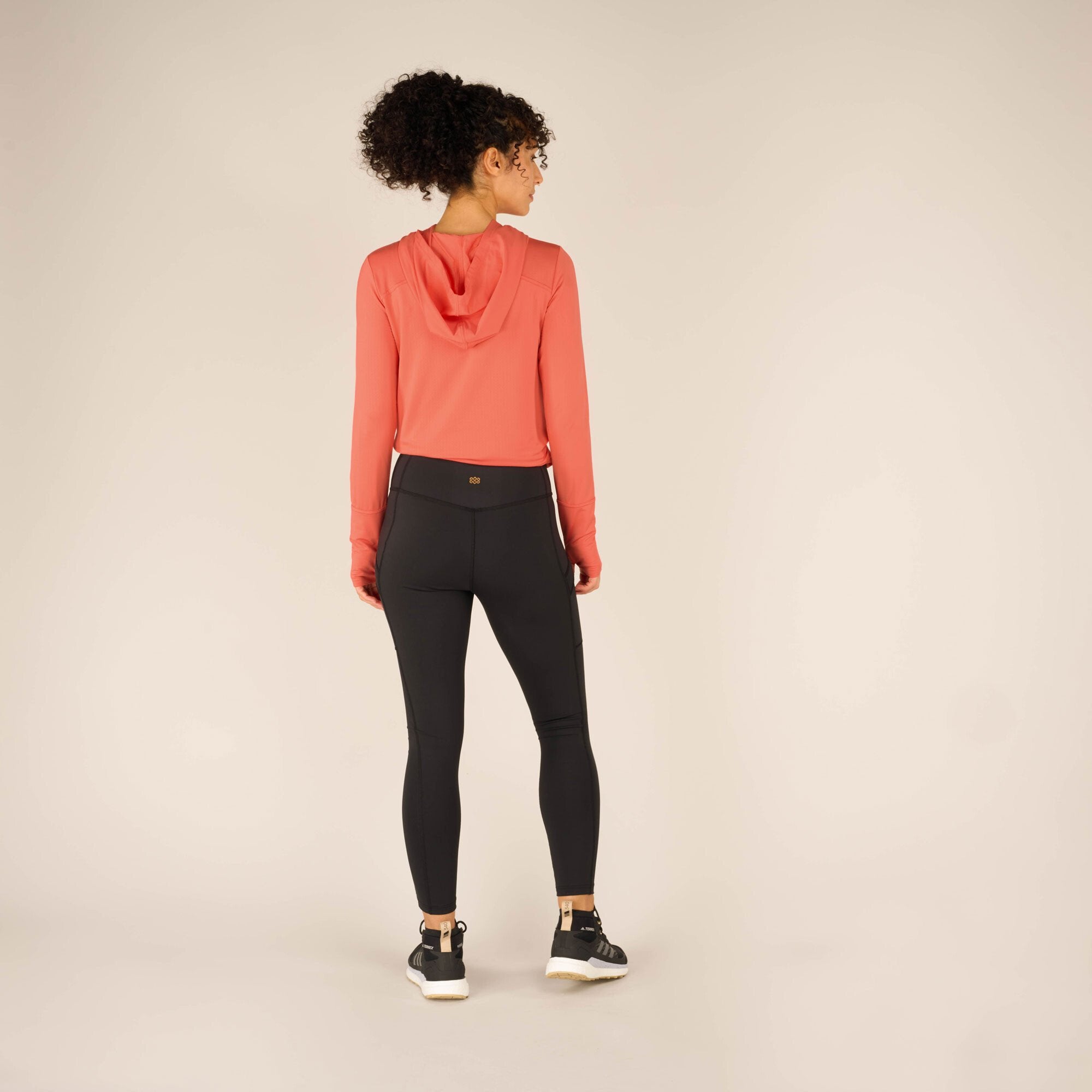 Back view of the model wearing the coral athletic top and Sherpa Adventure Gear Nisha Tight in Black. The leggings have a snug fit around the waist and hips with a small logo on the waistband. Vertical seams run down the back of the legs, enhancing the fitted silhouette. The coral top features a hood that drapes loosely. The model’s curly hair is tied in a high ponytail, and the black athletic shoes remain visible.