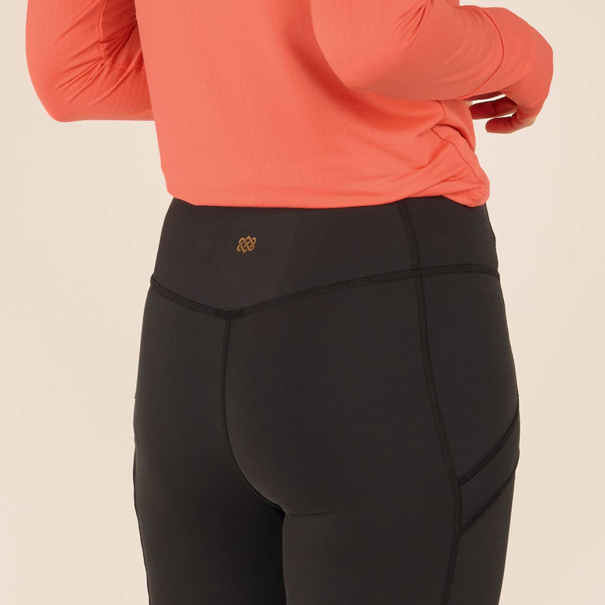 Close-up shot of the model’s lower back and waist, focusing on the Sherpa Adventure Gear Nisha Tight in Black. A small gold logo is visible at the center of the waistband. The stitching on the leggings runs diagonally, adding a structured look. The high waist sits just above the hips, offering a secure fit.