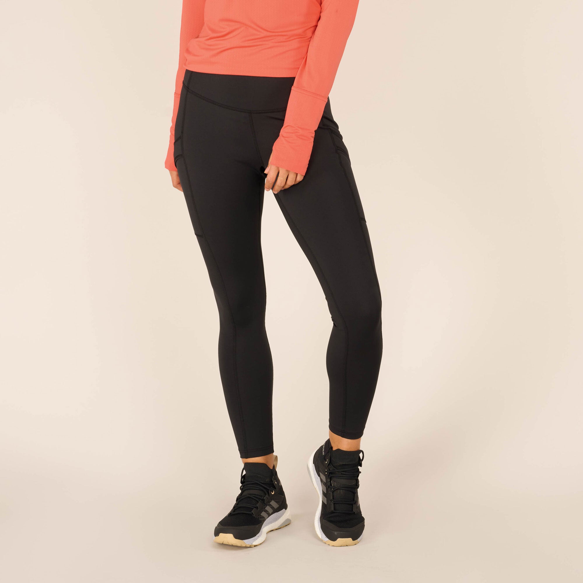 Front view of the model’s lower body, focusing on the Sherpa Adventure Gear Nisha Tight in Black. The leggings contour the thighs and calves, with seams highlighting the fit. The coral athletic top is partially visible, and the model’s stance showcases the leggings' flexibility and structure. The black athletic shoes with white soles complete the sporty look.