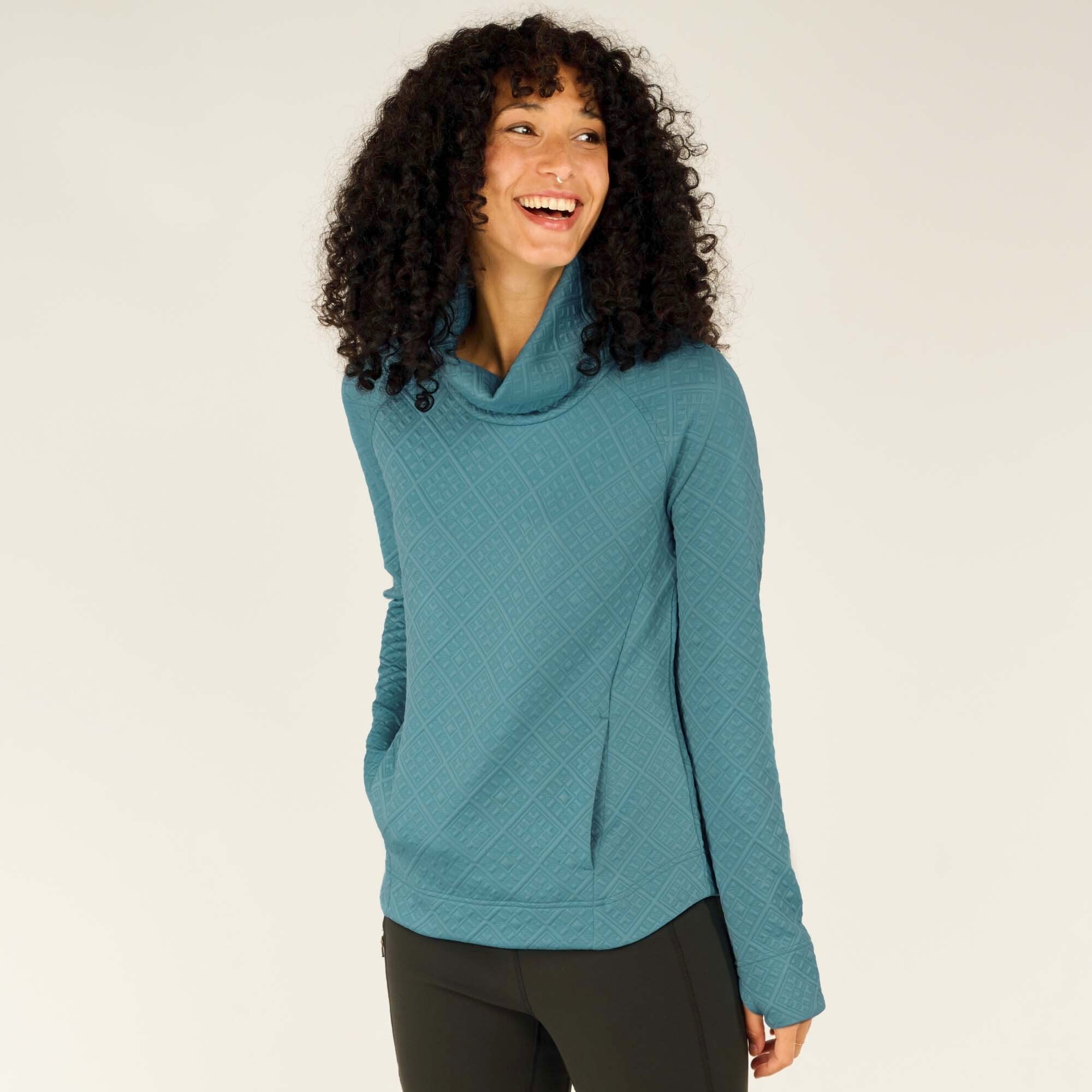 A young woman with curly black hair and a bright smile is modeling the Sherpa Adventure Gear Nyano Pullover in Blue. The pullover features a diamond quilted pattern, a cozy cowl neck, and long sleeves. She stands at a slight angle with one hand in her pocket, looking off to the side. The background is plain off-white.