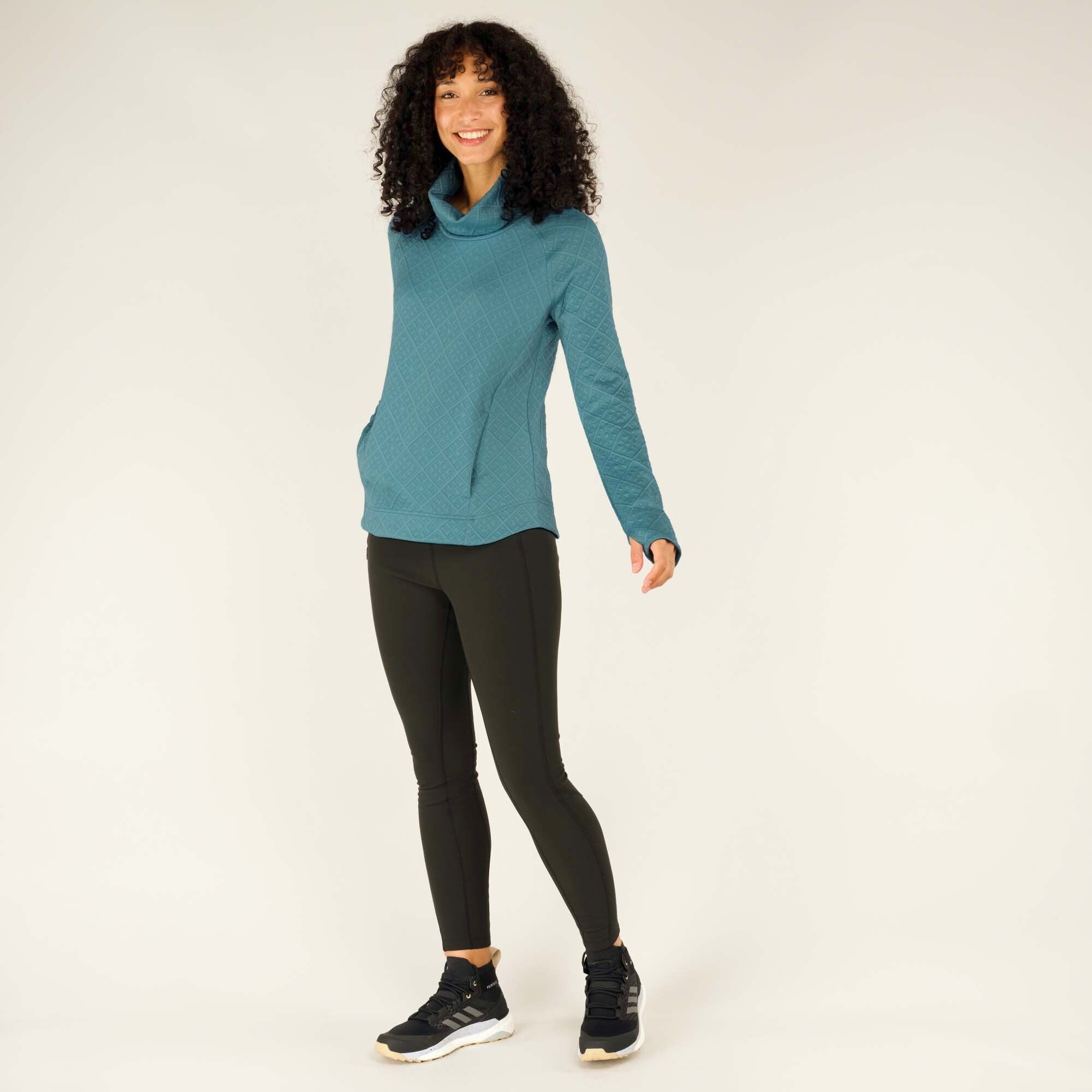 The same model is shown in a full-body shot, standing with her feet slightly apart. She wears the Sherpa Adventure Gear Nyano Pullover in Blue along with black leggings and black sneakers with white soles. She has a cheerful expression and a relaxed posture, highlighting the casual and comfortable fit of the pullover.