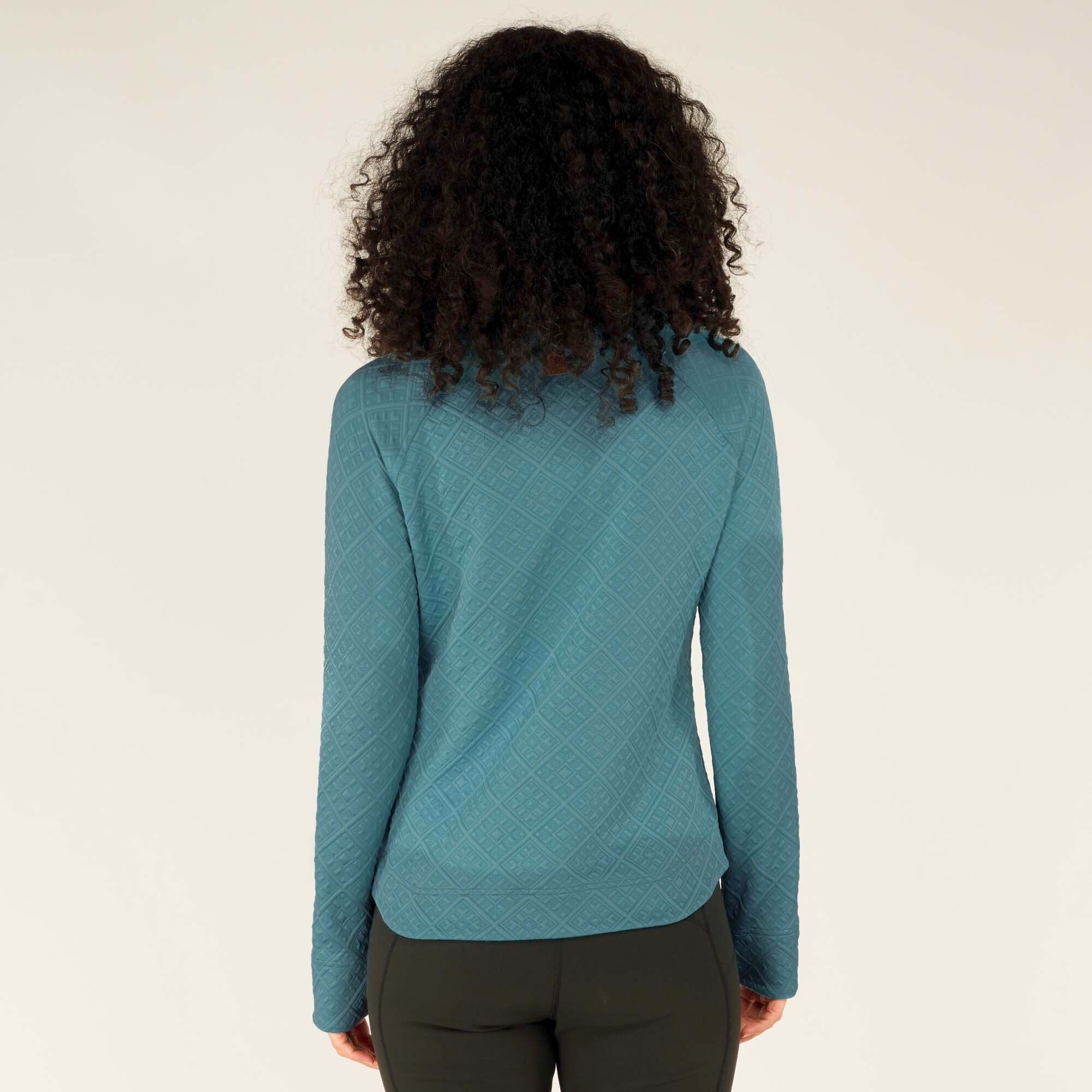 A back view of the model showcases the full length of the Sherpa Adventure Gear Nyano Pullover in Blue, with its textured diamond quilted design extending across the entire back. The slightly curved hemline and relaxed fit are visible. Her curly hair cascades down, partially covering the upper back of the pullover.