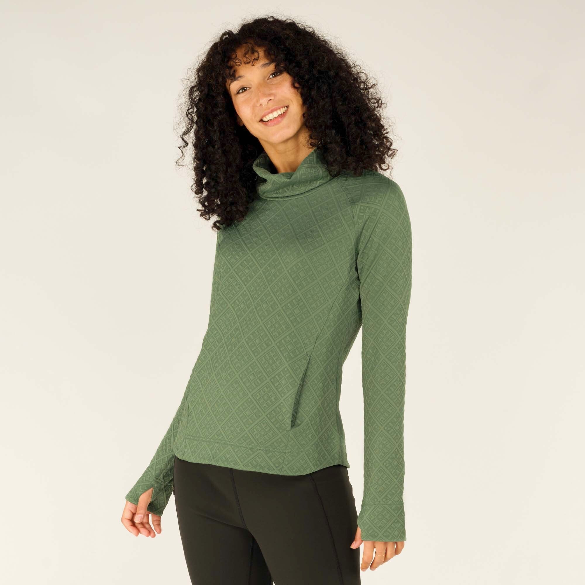 A woman with curly dark hair and a warm smile is modeling a fitted Sherpa Adventure Gear Nyano Pullover in Green with a quilted diamond pattern. The pullover features a loose cowl neck, long sleeves with thumbholes, and two angled front pockets. She is wearing black leggings and is slightly angled to her left, looking at the camera with a relaxed expression.