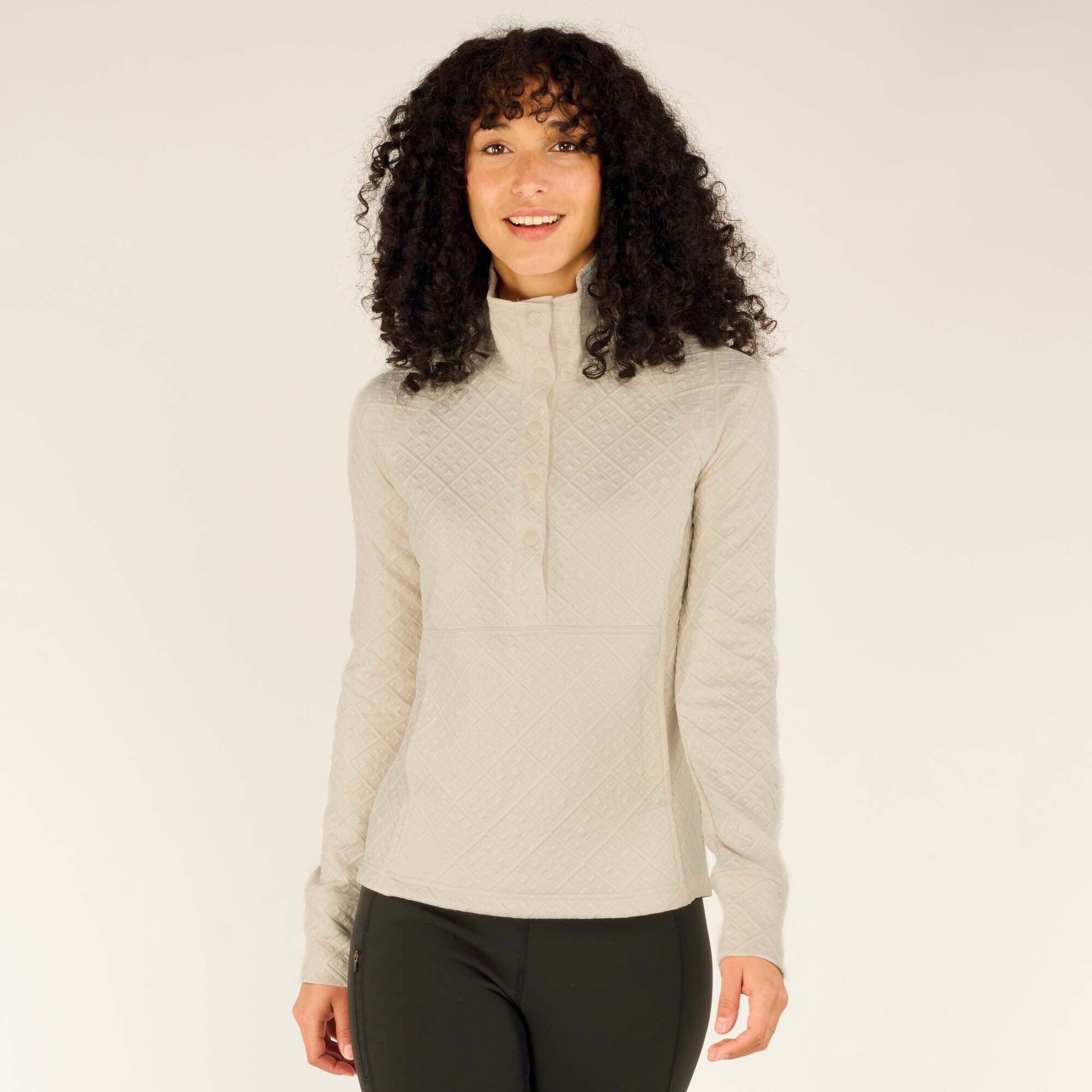 The image showcases a woman wearing a Sherpa Adventure Gear Nyano Snap Mock Neck in Green with a high mock-neck collar featuring snap buttons down the front. The textured fabric displays a diamond pattern, offering a stylish and cozy look. She pairs the top with black leggings and stands facing forward, smiling softly.