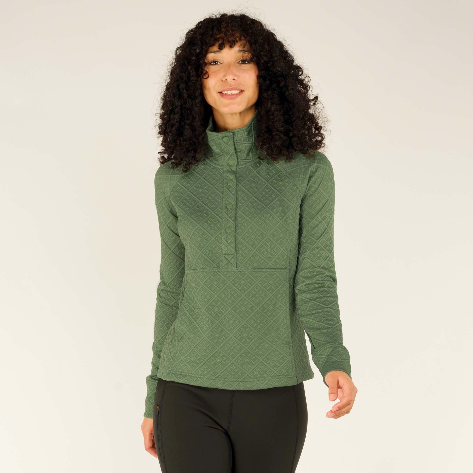 A woman with curly dark hair and a smile stands facing the camera wearing a Sherpa Adventure Gear Nyano Snap Mock Neck in Green with a diamond pattern. The pullover has a front snap closure at the collar, and the fitted design accentuates her shape. She pairs it with black leggings, standing in a relaxed posture with one arm slightly extended.