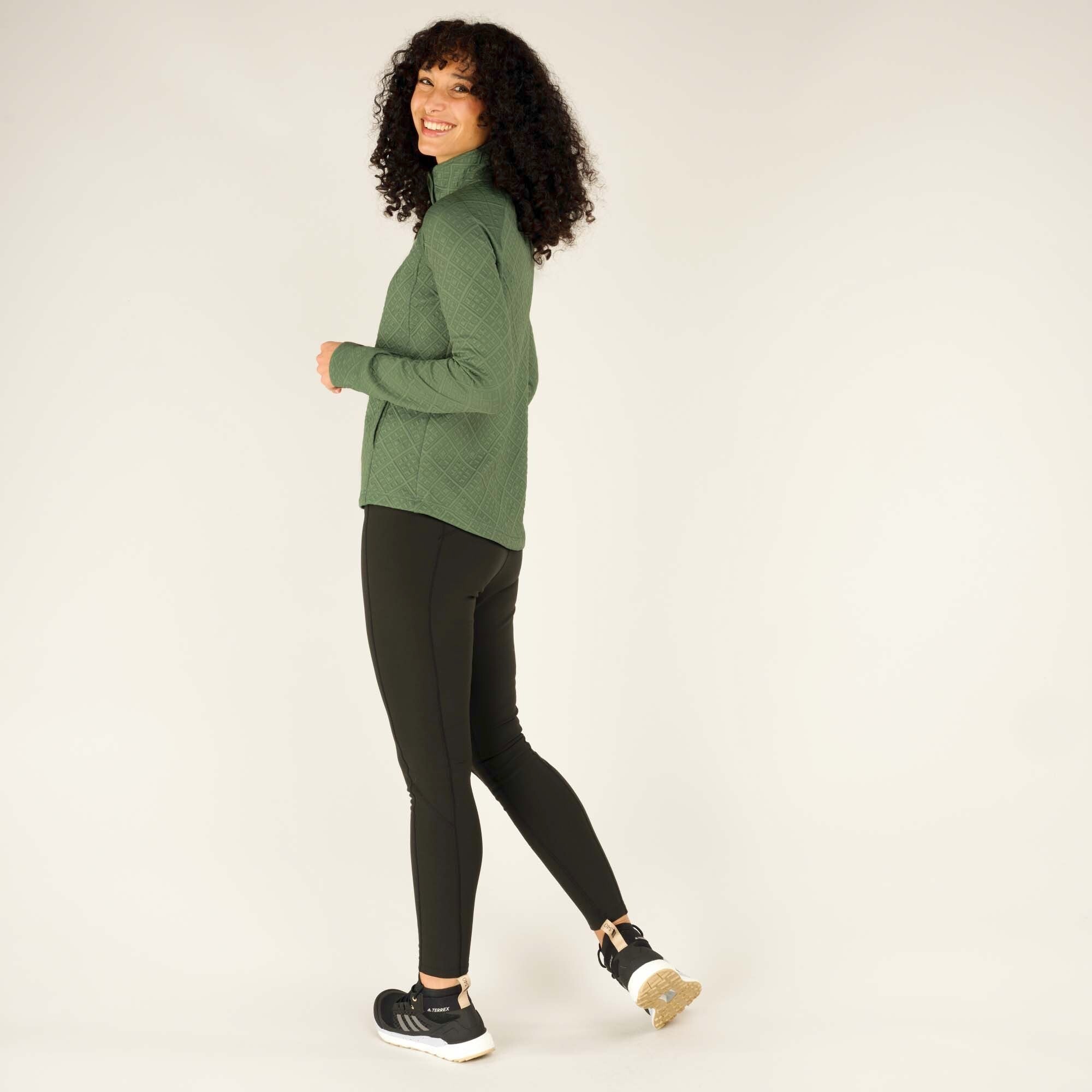 A side profile view of the woman wearing the same Sherpa Adventure Gear Nyano Snap Mock Neck in Green, walking slightly forward with a cheerful expression. Her curly hair frames her face, and she looks towards the camera while smiling. The pullover's fit and textured fabric are visible, and the zipped pockets add a practical touch to the design.