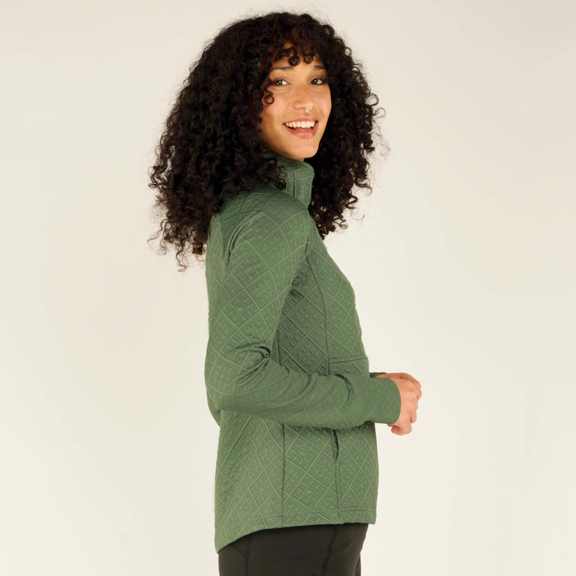 A closer side angle of the woman in the Sherpa Adventure Gear Nyano Snap Mock Neck in Green, with her hands tucked into the pockets. She is looking over her shoulder with a bright smile. The quilted diamond pattern and fitted sleeves are highlighted in this shot, emphasizing the textured details of the top.