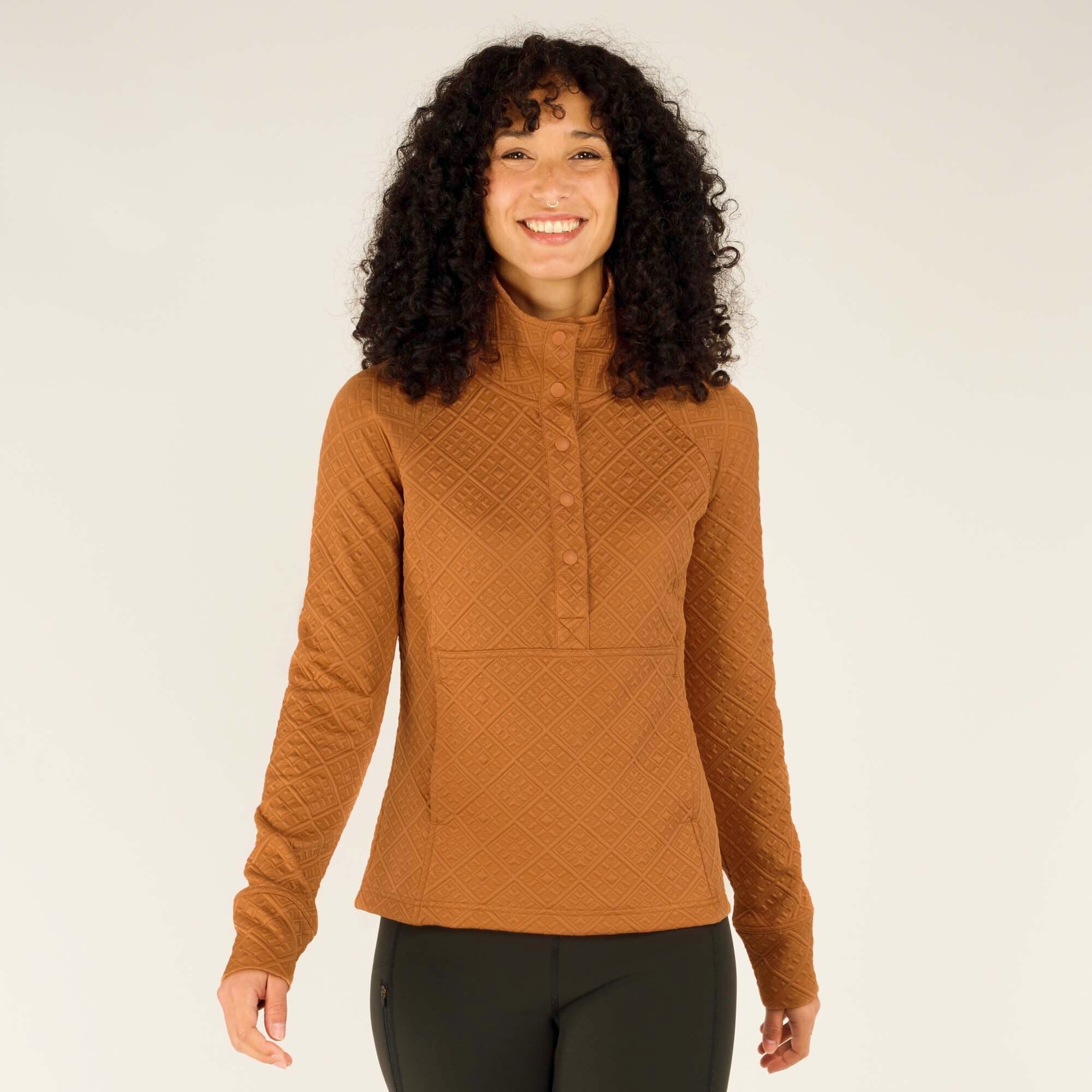 A woman with curly dark hair and a smile stands facing the camera, wearing a textured Sherpa Adventure Gear Nyano Snap Mock Neck in Orange with a mock neck and snap button closure. The top features a diamond quilted pattern throughout, long sleeves, and a slightly relaxed fit. She is also wearing dark green leggings.