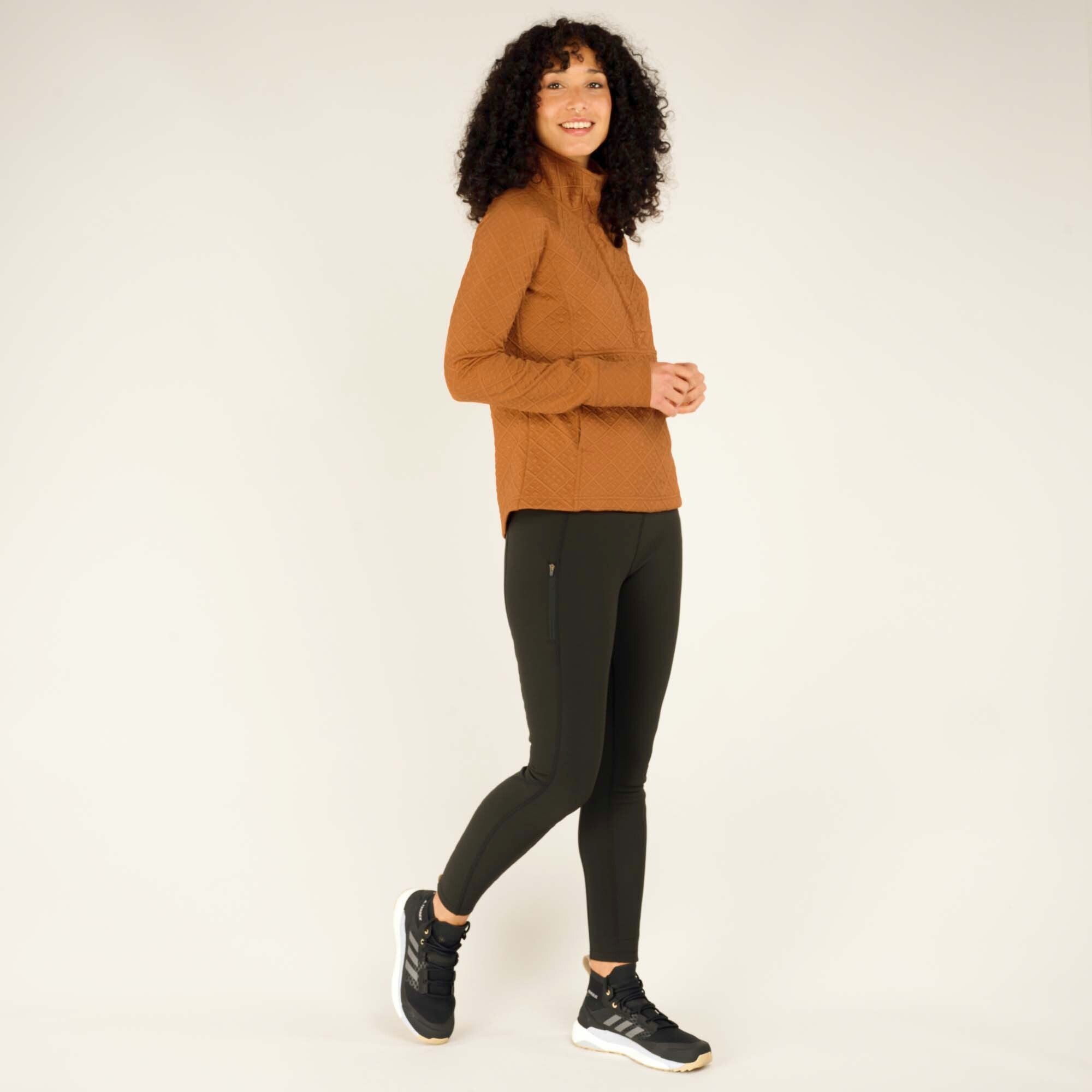 The same woman is shown in a side view, walking slightly forward with one foot raised. The Sherpa Adventure Gear Nyano Snap Mock Neck in Orange fits comfortably around her torso, with the hem curving slightly lower at the back. The mock neck stands up neatly, and the front snaps are partially visible. The diamond texture pattern covers the pullover entirely, and her hands are relaxed at her sides.