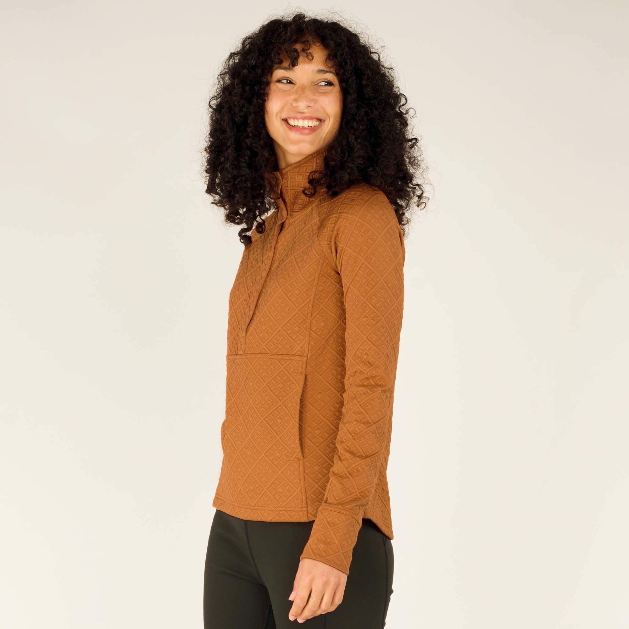 The woman is turned slightly toward the camera, showing the Sherpa Adventure Gear Nyano Snap Mock Neck in Orange’s side seam and part of the front snap closure. She is smiling broadly and her curly hair frames her face. The pullover’s fit is relaxed, with room for movement, and the diamond pattern is clearly visible across the chest, sleeves, and side.