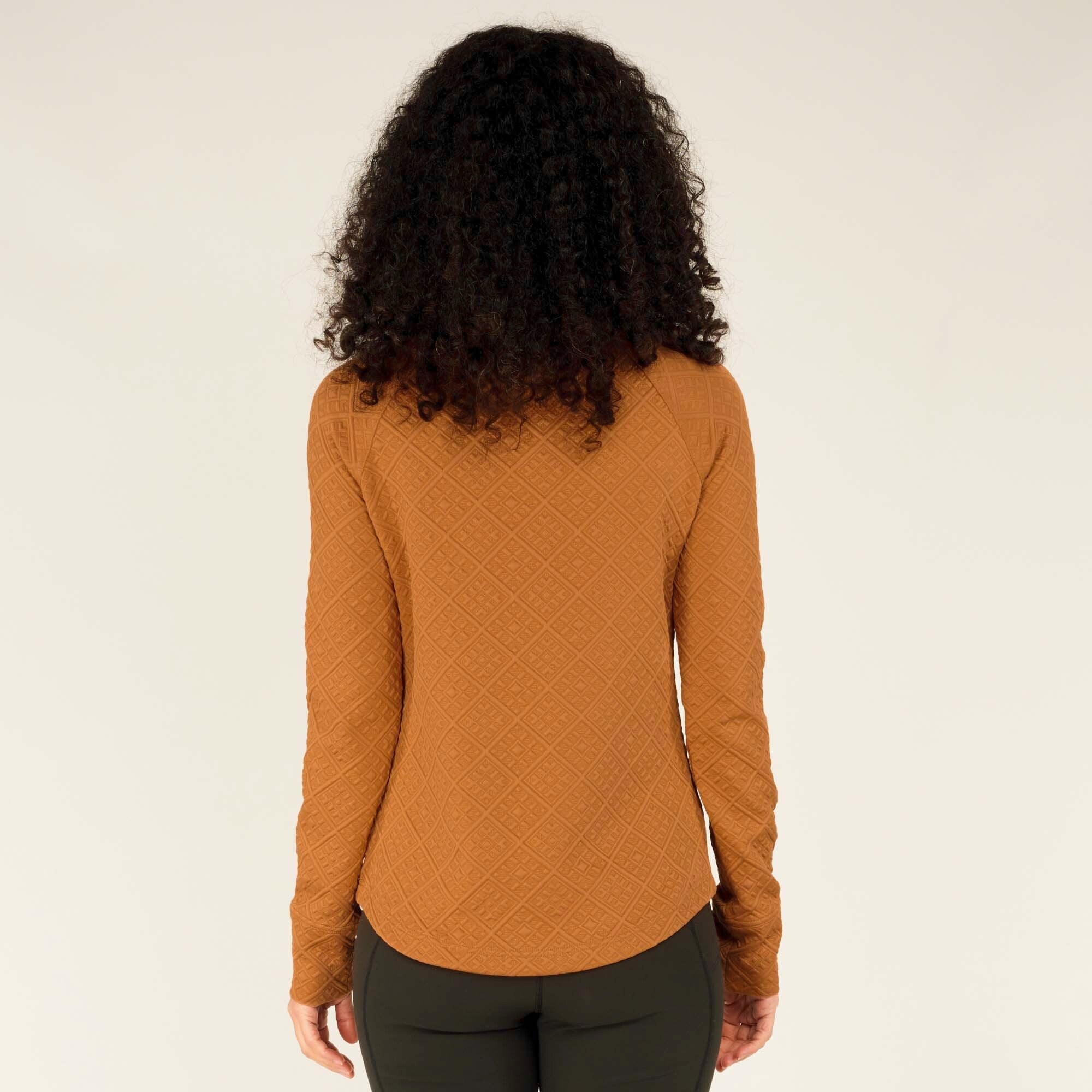 The woman stands with her back to the camera, arms relaxed at her sides, showing the full back view of the Sherpa Adventure Gear Nyano Snap Mock Neck in Orange. The diamond quilted texture continues across the entire back. The hem dips slightly lower at the back for extra coverage, and the fabric appears soft and flexible.