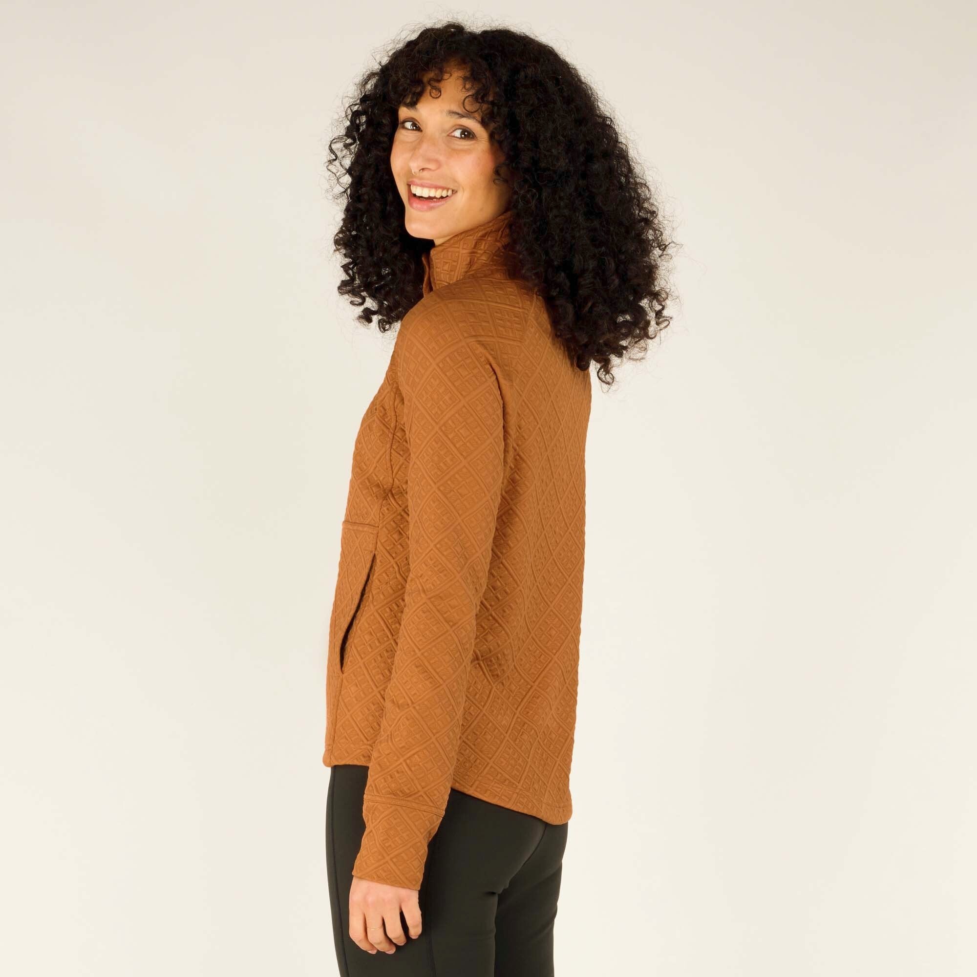 In this final image, the woman stands in a relaxed side pose, smiling while looking toward the camera. Her left hand is tucked into the front kangaroo-style pocket of the Sherpa Adventure Gear Nyano Snap Mock Neck in Orange, which blends seamlessly into the diamond texture. The mock neck and snap closure add a functional and stylish touch to the cozy design.