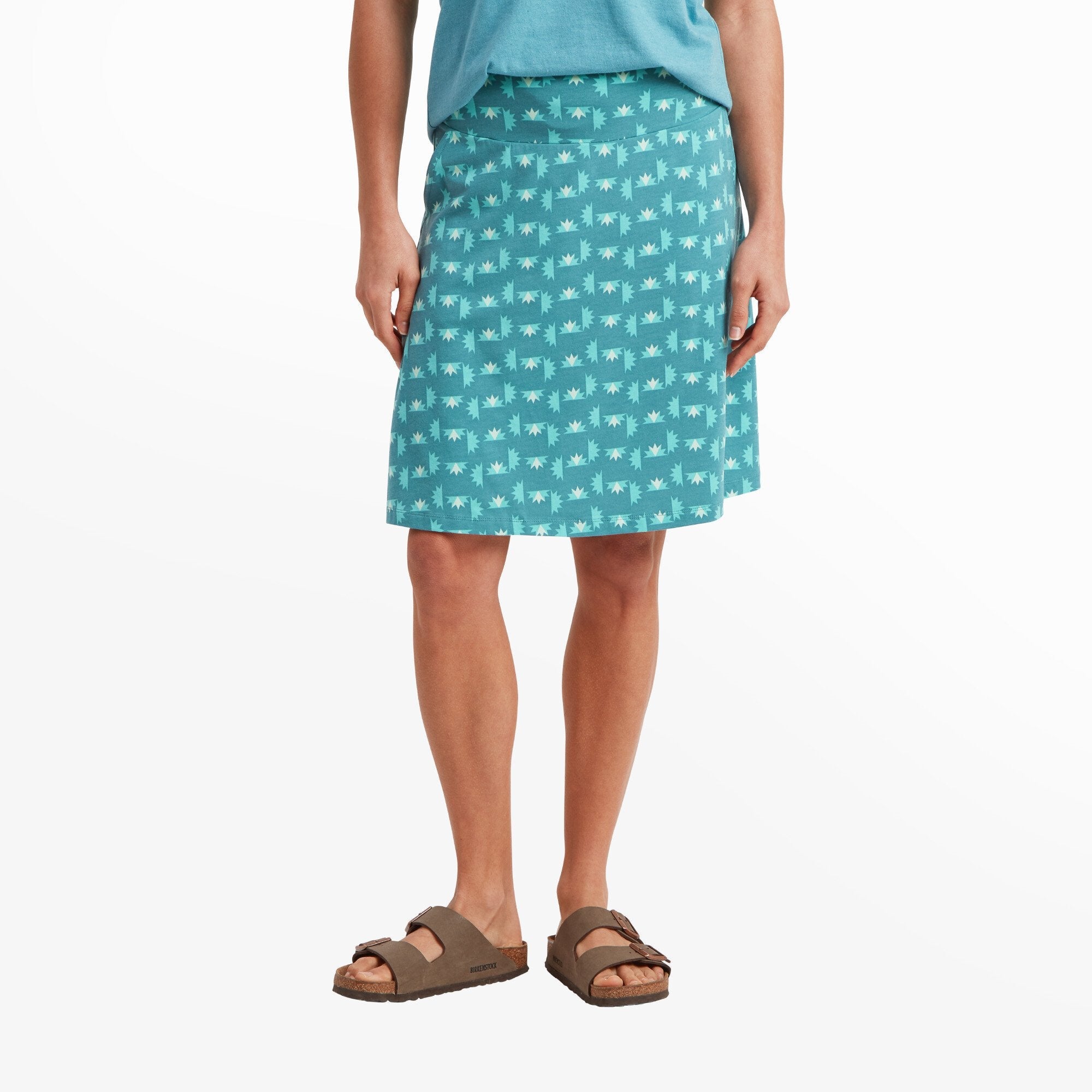 A woman stands facing forward wearing the Sherpa Adventure Gear Padma Pull On Skirt in Blue with a playful, geometric print of light blue and white shapes resembling stylized flowers. The skirt falls just above the knees and has a flat, wide waistband for comfort. She pairs the skirt with a matching blue tank top tucked in slightly at the waist and wears brown sandals.