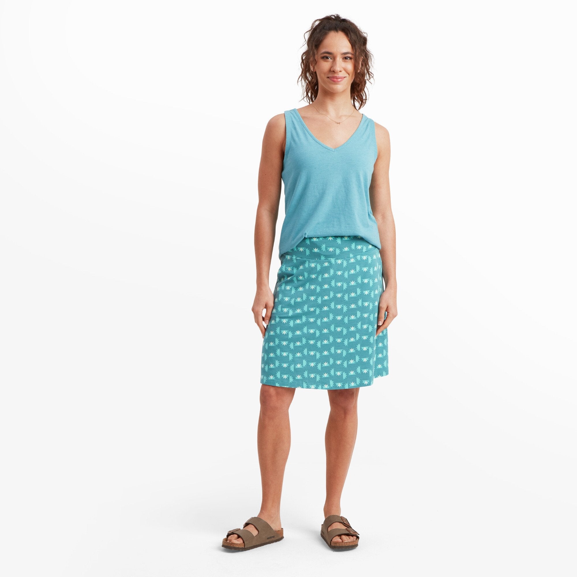 Full-body shot of the same woman modeling the Sherpa Adventure Gear Padma Pull On Skirt in Blue. She stands relaxed, smiling slightly, with one hand at her side and the other slightly bent. The skirt’s print and color contrast nicely with the solid blue tank top she’s wearing. The look is casual and summery, with comfortable sandals completing the outfit.