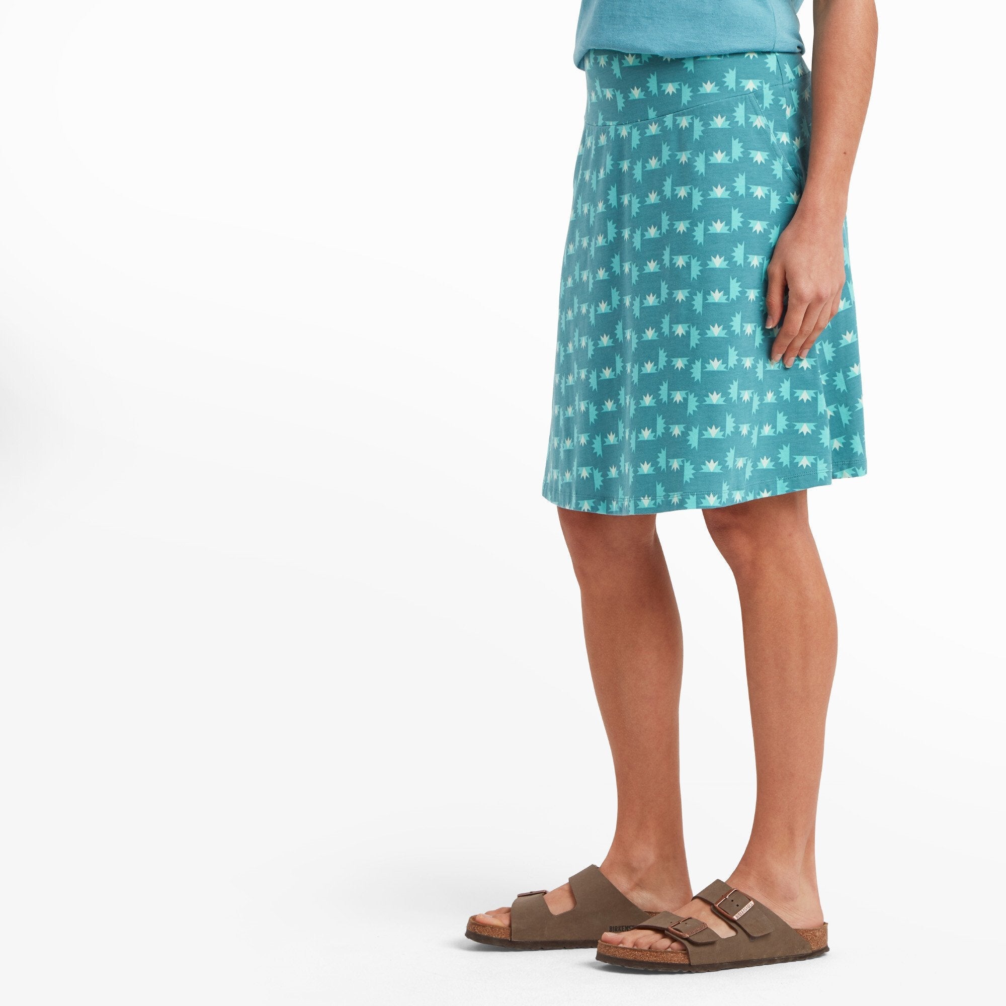 A side view of the woman wearing the Sherpa Adventure Gear Padma Pull On Skirt in Blue, showing its gentle A-line silhouette. The fabric drapes smoothly over her hips, and the playful print continues uninterrupted around the skirt. Her left hand rests along her side, showcasing the smooth waistband.