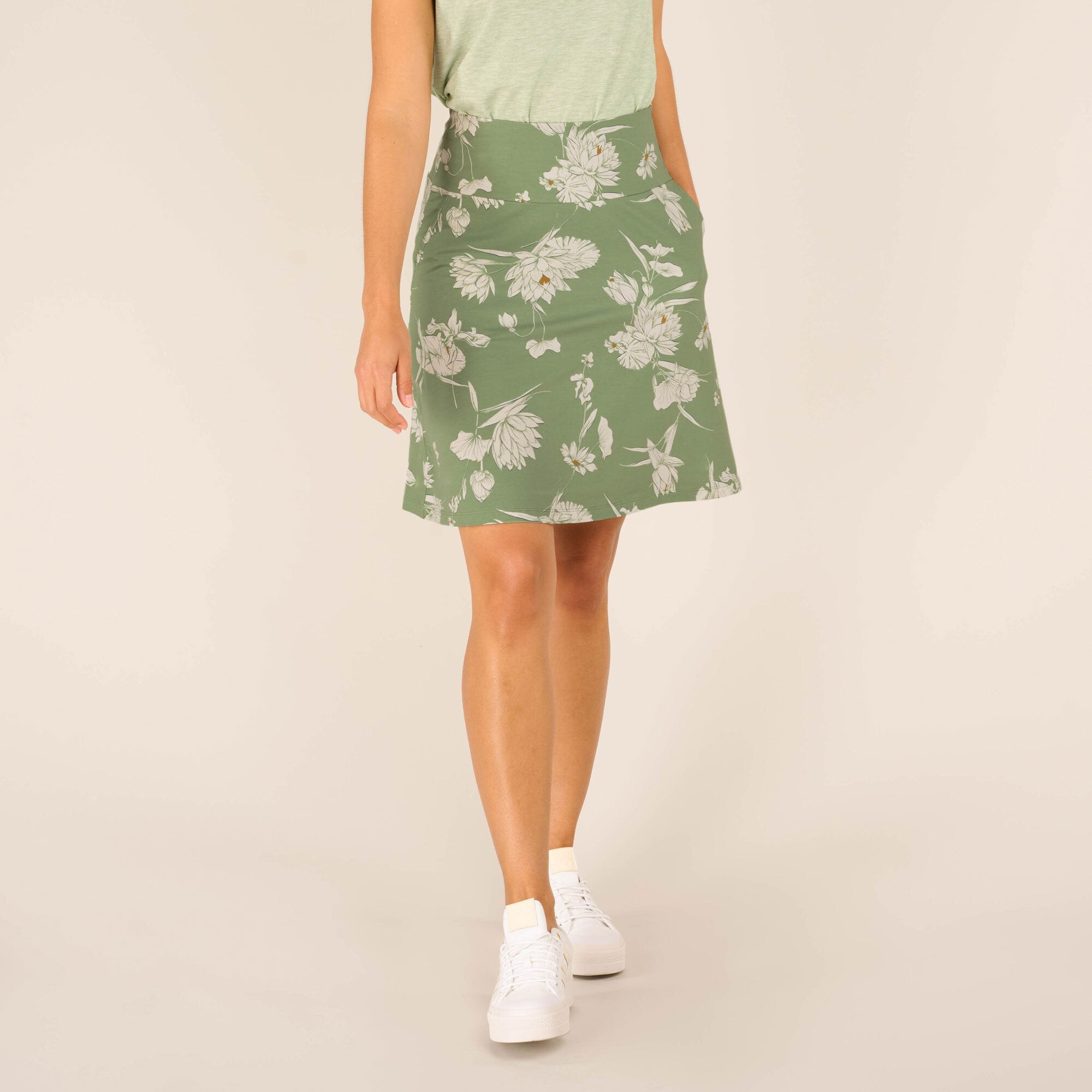 A close-up front view of a Sherpa Adventure Gear Padma Pull On Skirt in Green. The model stands with one leg slightly forward, showcasing the elegant A-line silhouette. The skirt features delicate white floral patterns and side pockets, paired with casual white trainers and a light green top.