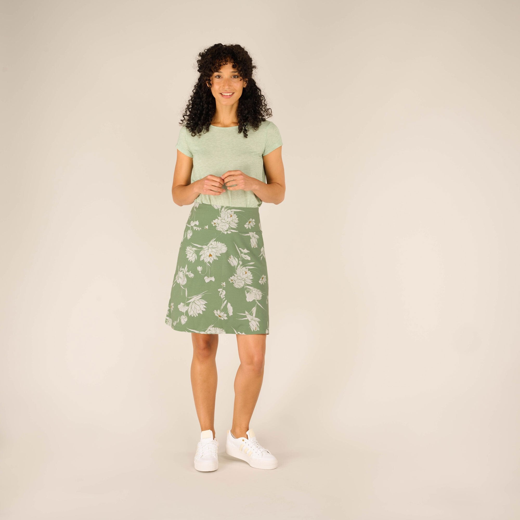 The model is fully visible wearing the Sherpa Adventure Gear Padma Pull On Skirt in Green paired with a matching light green short-sleeve top. She smiles while standing confidently, hands clasped together in front, highlighting the relaxed fit of the skirt.