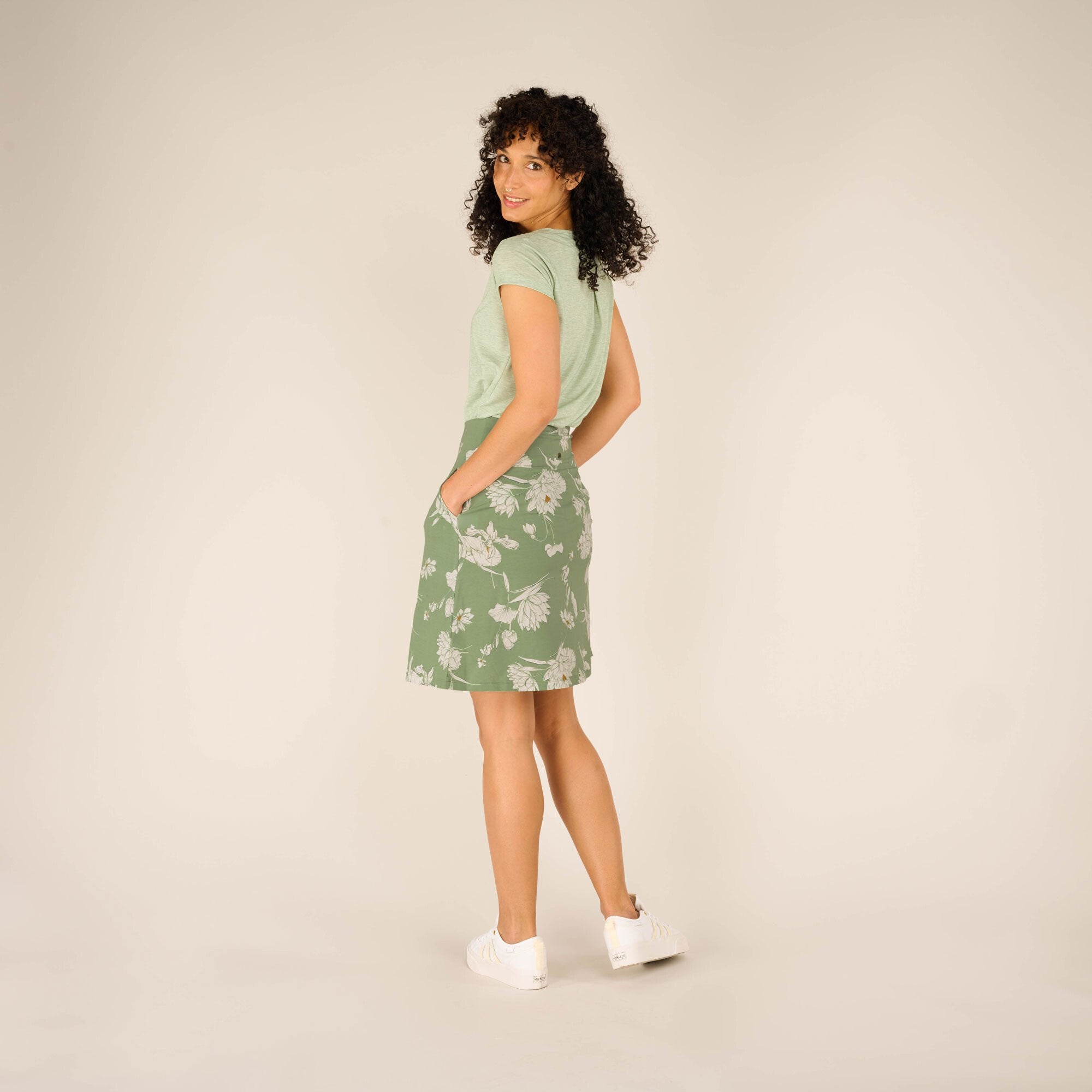 A side profile view of the model wearing the Sherpa Adventure Gear Padma Pull On Skirt in Green. Her hands are tucked into the skirt’s pockets, emphasising the practicality of the design. The floral print flows seamlessly across the soft green fabric, and the look is complemented by white trainers.