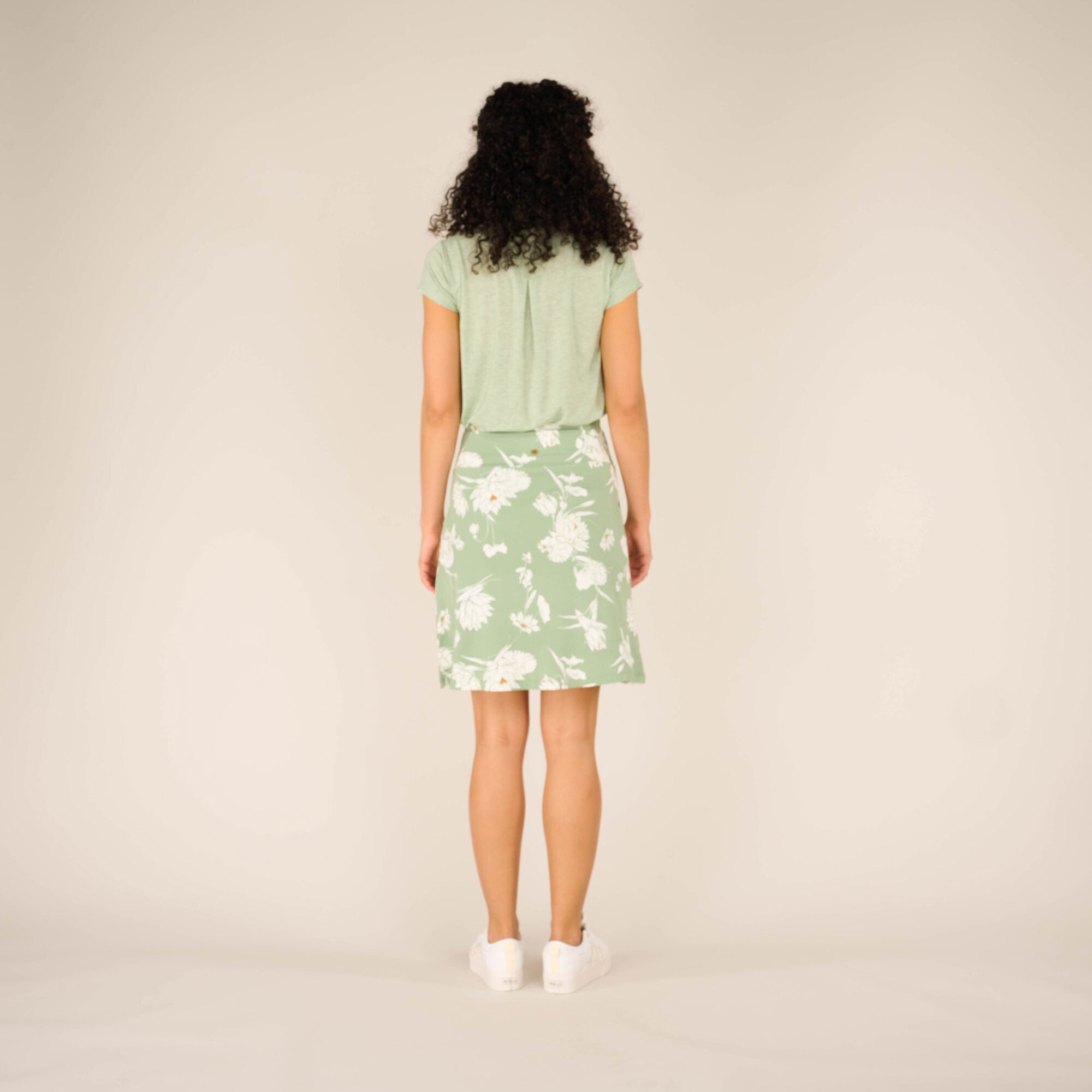 The back view of the Sherpa Adventure Gear Padma Pull On Skirt in Green. The model faces away, showcasing the gentle curve of the waistband and the relaxed hemline that falls just above the knee. Her casual white trainers complete the outfit.