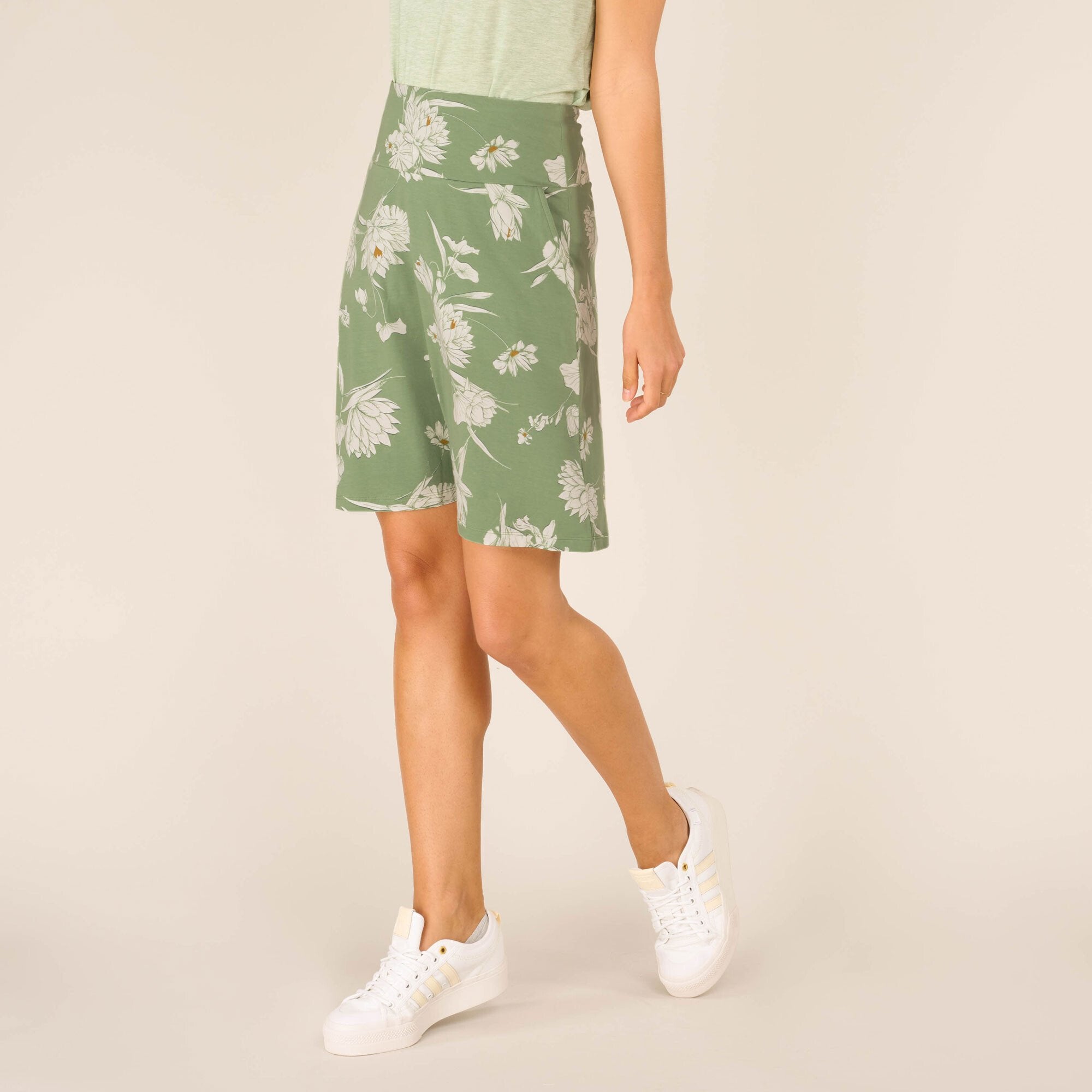 A diagonal front view of the model in the Sherpa Adventure Gear Padma Pull On Skirt in Green. Her posture is relaxed with one leg slightly forward, emphasising the skirt’s flow and side-pocket detailing. The soft floral print appears vibrant against the green background.