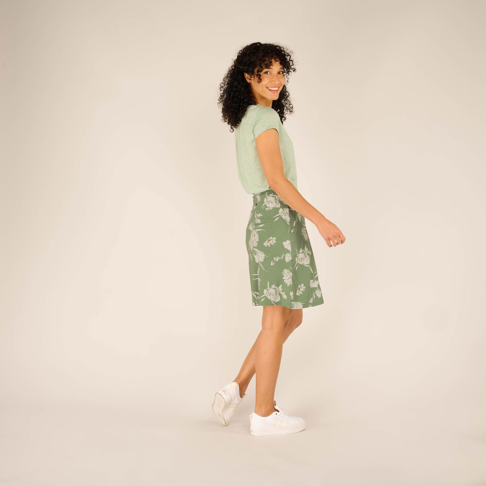 A playful side-back view of the model wearing the Sherpa Adventure Gear Padma Pull On Skirt in Green as she looks over her shoulder with a smile. The green floral-printed skirt flows naturally, complementing her movement. The relaxed and versatile design is paired with casual white trainers for a breezy summer look.