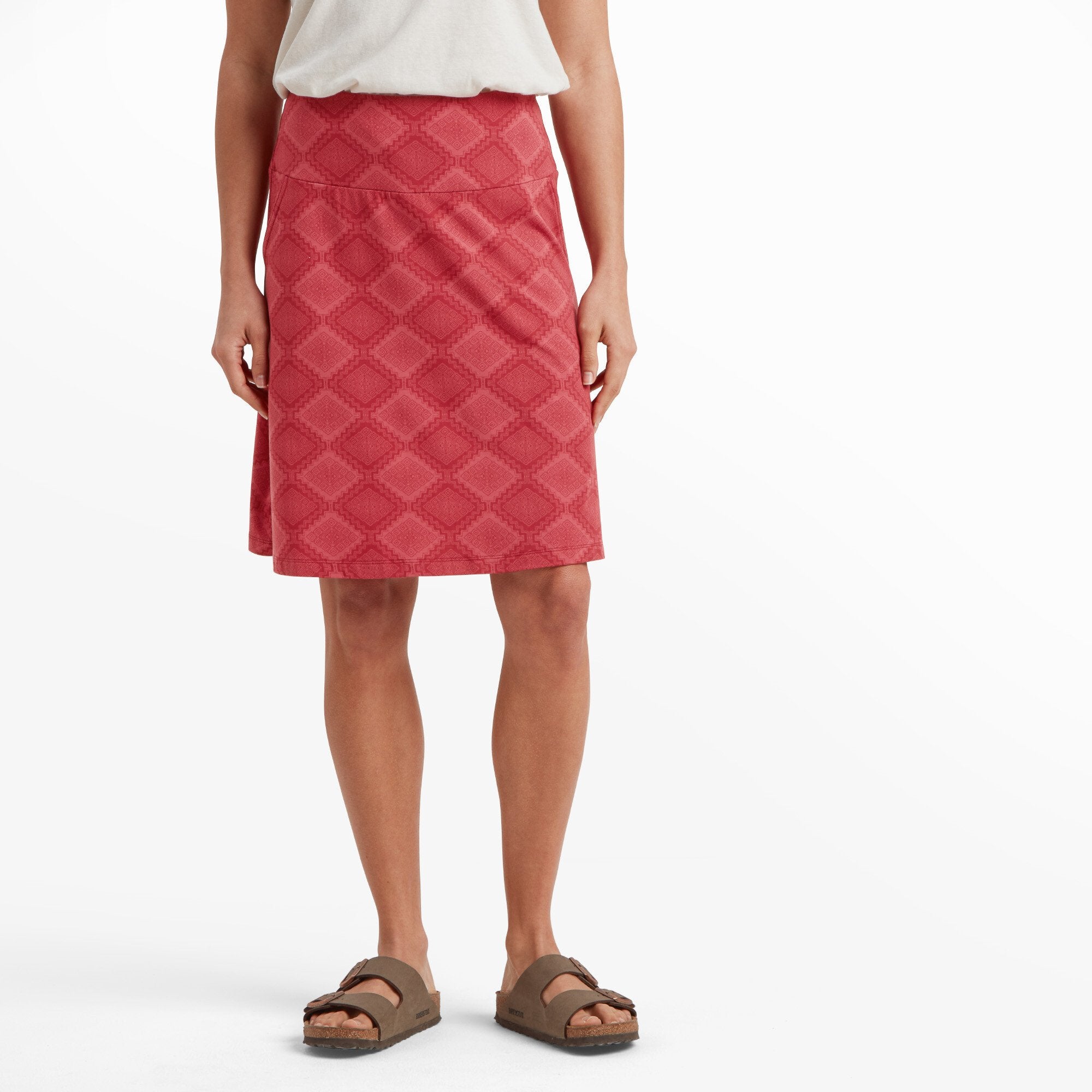 A close-up view of a woman's lower body showcasing a Sherpa Adventure Gear Padma Pull On Skirt in Red with an intricate geometric pattern. The skirt is knee-length, fitted at the waist, and flares slightly towards the bottom. She is wearing a tucked-in white top and brown open-toe sandals.