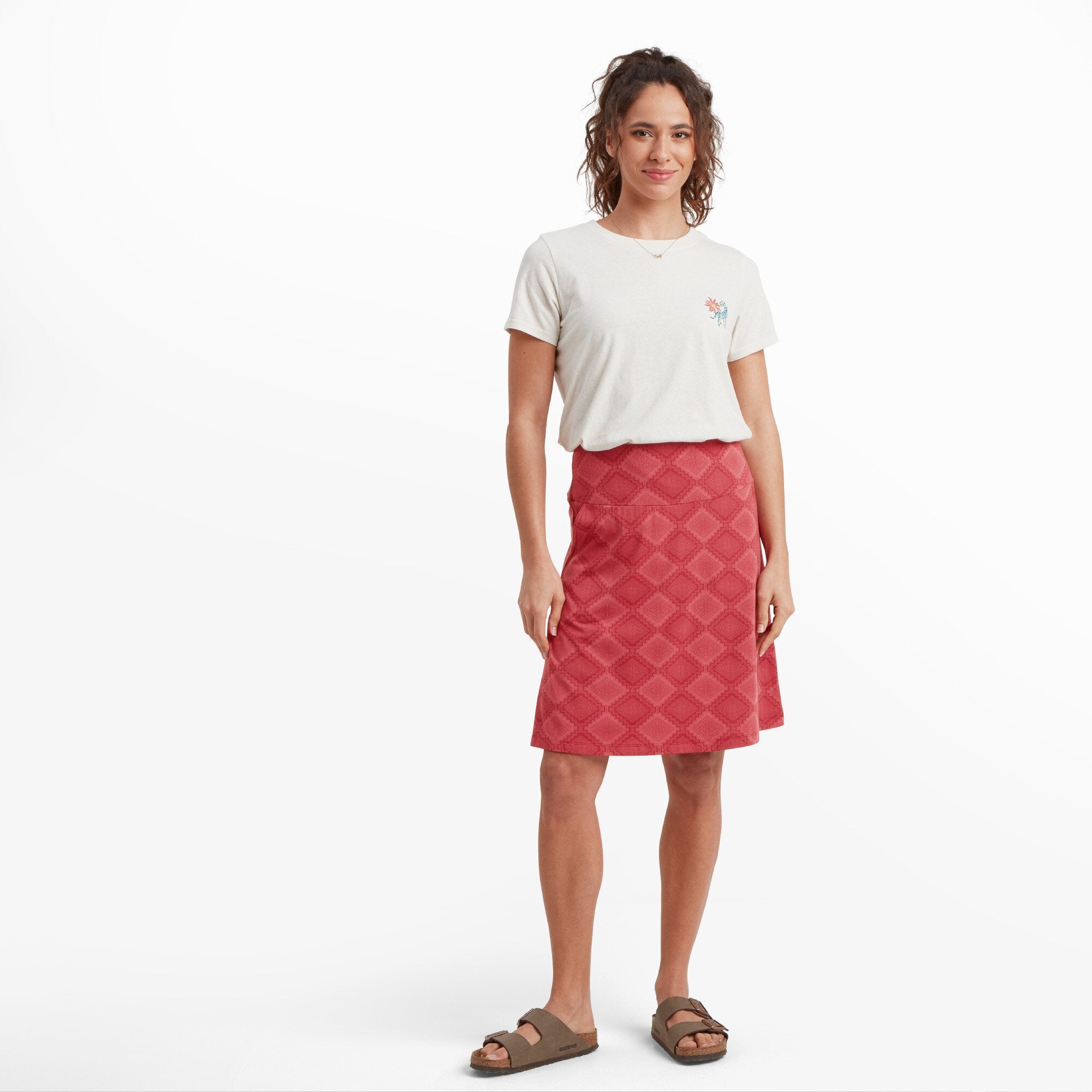 A smiling woman poses in a knee-length Sherpa Adventure Gear Padma Pull On Skirt in Red with an intricate geometric pattern, paired with a white short-sleeve t-shirt featuring a small floral embroidery on the chest. She is wearing brown sandals, and the outfit is set against a plain white background.