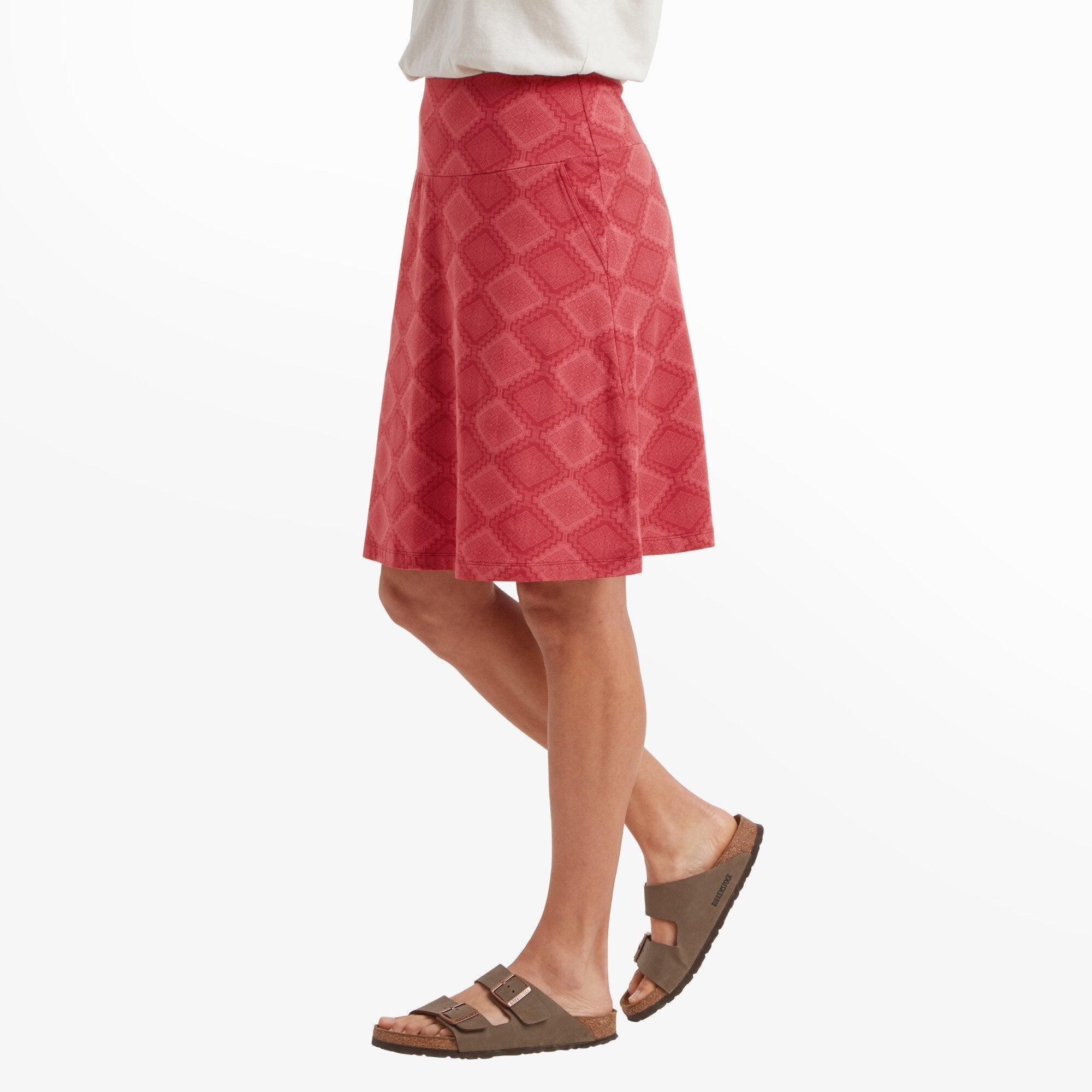 A woman is captured from the side, highlighting the A-line silhouette of the Sherpa Adventure Gear Padma Pull On Skirt in Red. The high-waisted design sits comfortably, and the skirt falls just above the knees. She is wearing a relaxed white top and brown sandals.