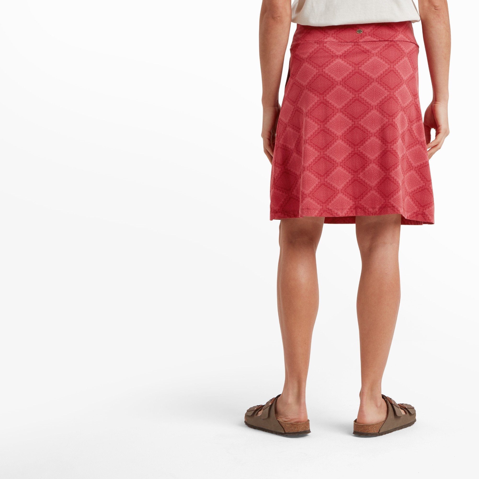 A woman is standing with her back facing the camera, displaying the back design of the Sherpa Adventure Gear Padma Pull On Skirt in Red. A small button detail is visible near the waistband, and the geometric pattern continues seamlessly. The skirt slightly flares at the bottom, and she pairs it with a white top and brown sandals.