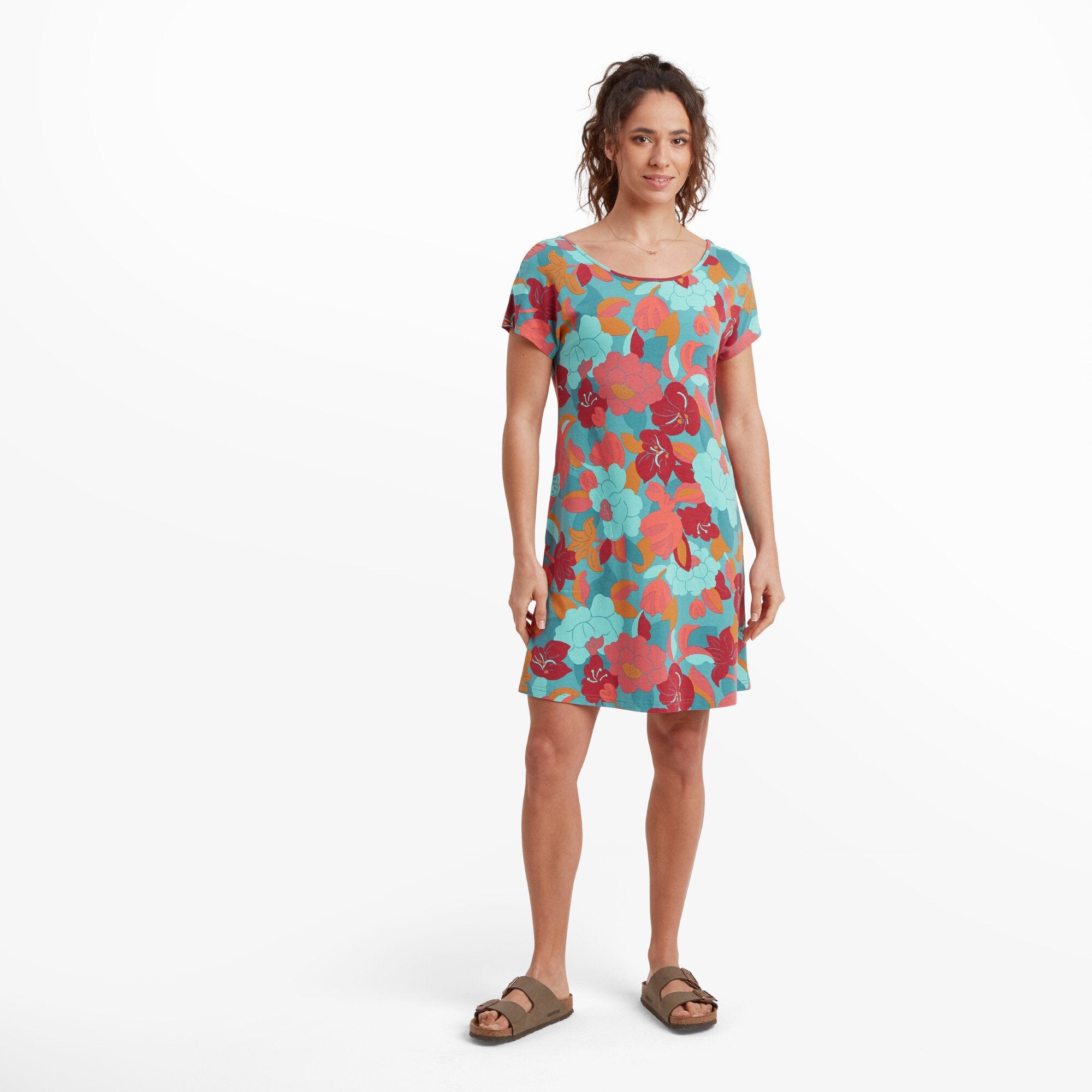 A woman is wearing a Sherpa Adventure Gear Padma Tie Back Dress in Blue with a relaxed fit. The dress features a scoop neckline, short sleeves, and a vibrant pattern of blue, red, and orange flowers. She is standing with her arms at her sides, wearing casual brown sandals. The background is white, emphasising the bright floral design.