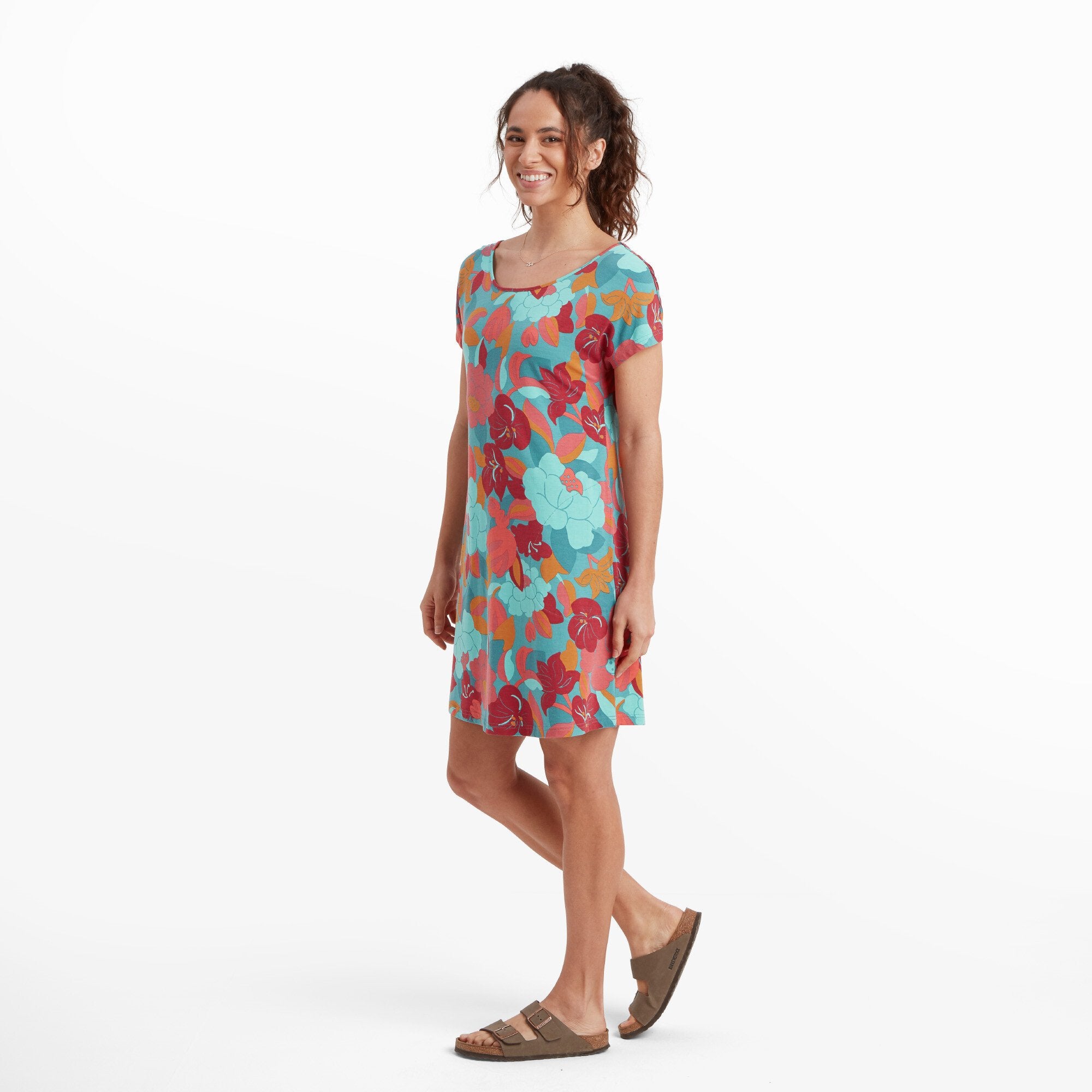 The same woman is shown in a side view, highlighting the silhouette of the Sherpa Adventure Gear Padma Tie Back Dress in Blue. The short sleeves and the dress's light, flowy material are visible. She is smiling and standing in a relaxed posture with one leg slightly bent. The brown sandals complement the dress's vibrant colors.