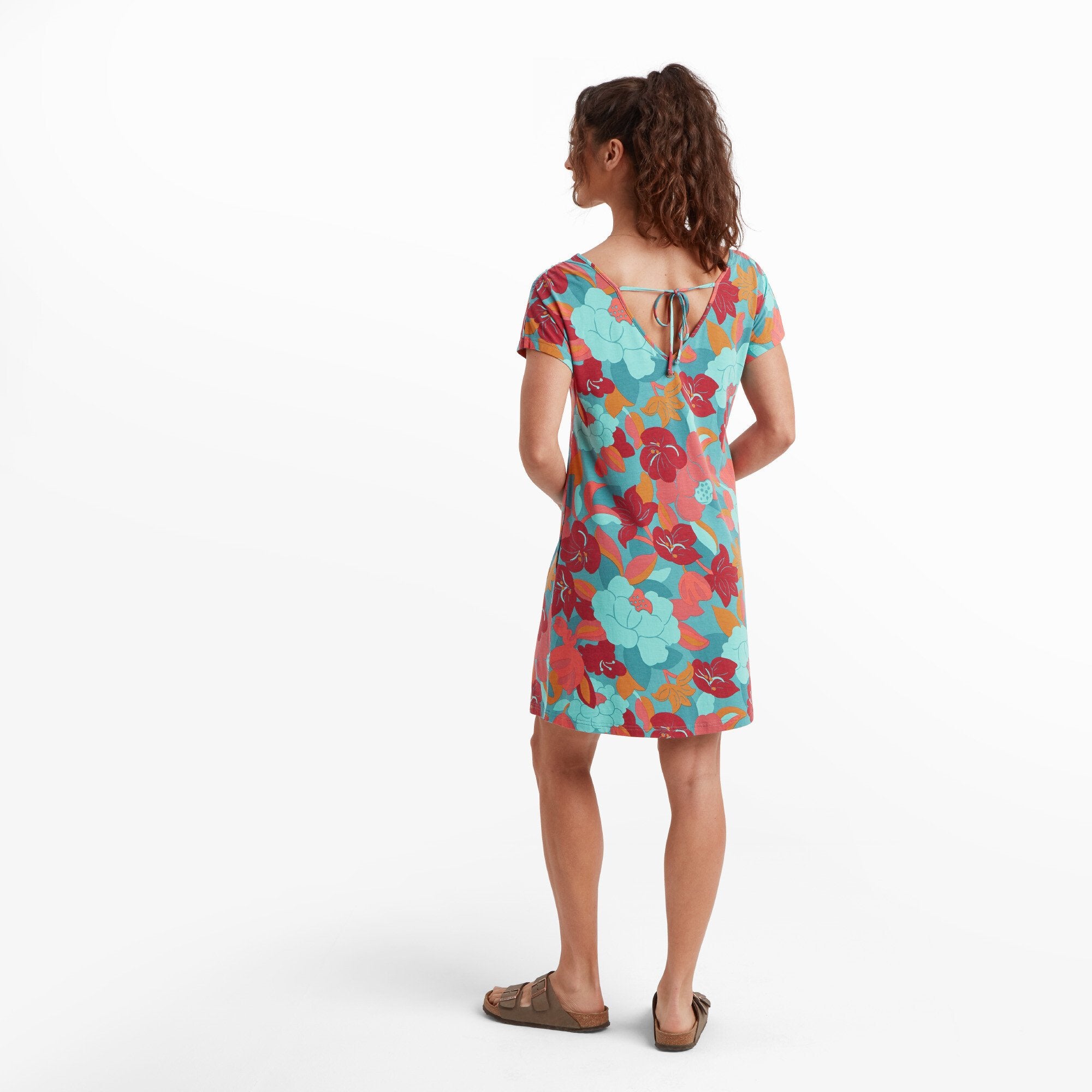 A back view of the woman wearing the Sherpa Adventure Gear Padma Tie Back Dress in Blue. The dress has a V-shaped back with a tie detail at the neckline, adding an elegant touch. The colorful floral pattern continues across the back. Her hair is tied up, showcasing the dress's design details.