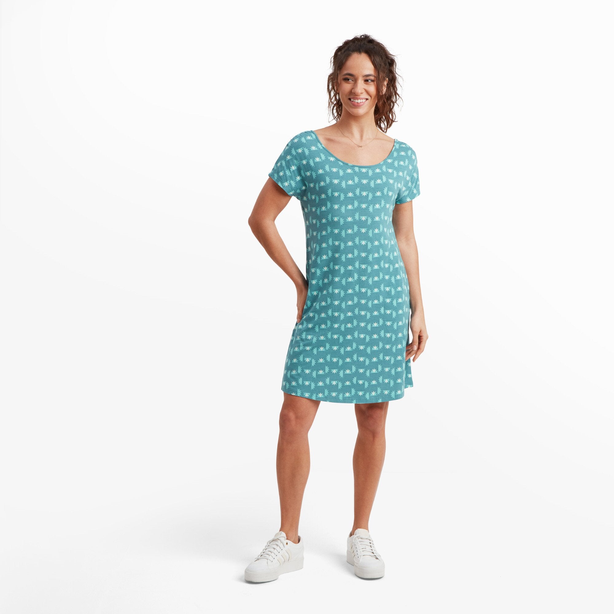 A woman is wearing a Sherpa Adventure Gear Padma Tie Back Dress in Blue with a subtle white geometric pattern. The dress features a scoop neckline and short sleeves, falling just above the knees. She is smiling, standing casually with one hand on her hip, and wearing white trainers.