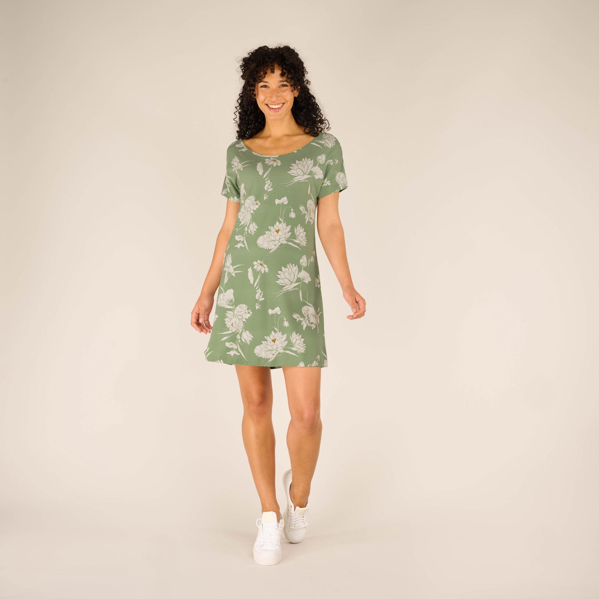 A smiling woman models a Sherpa Adventure Gear Padma Tie Back Dress in Green with short sleeves and a rounded neckline. She is standing and facing forward, showcasing the lightweight, flowy fabric of the dress. Her white trainers add a casual vibe, and the neutral background ensures the focus stays on the dress.