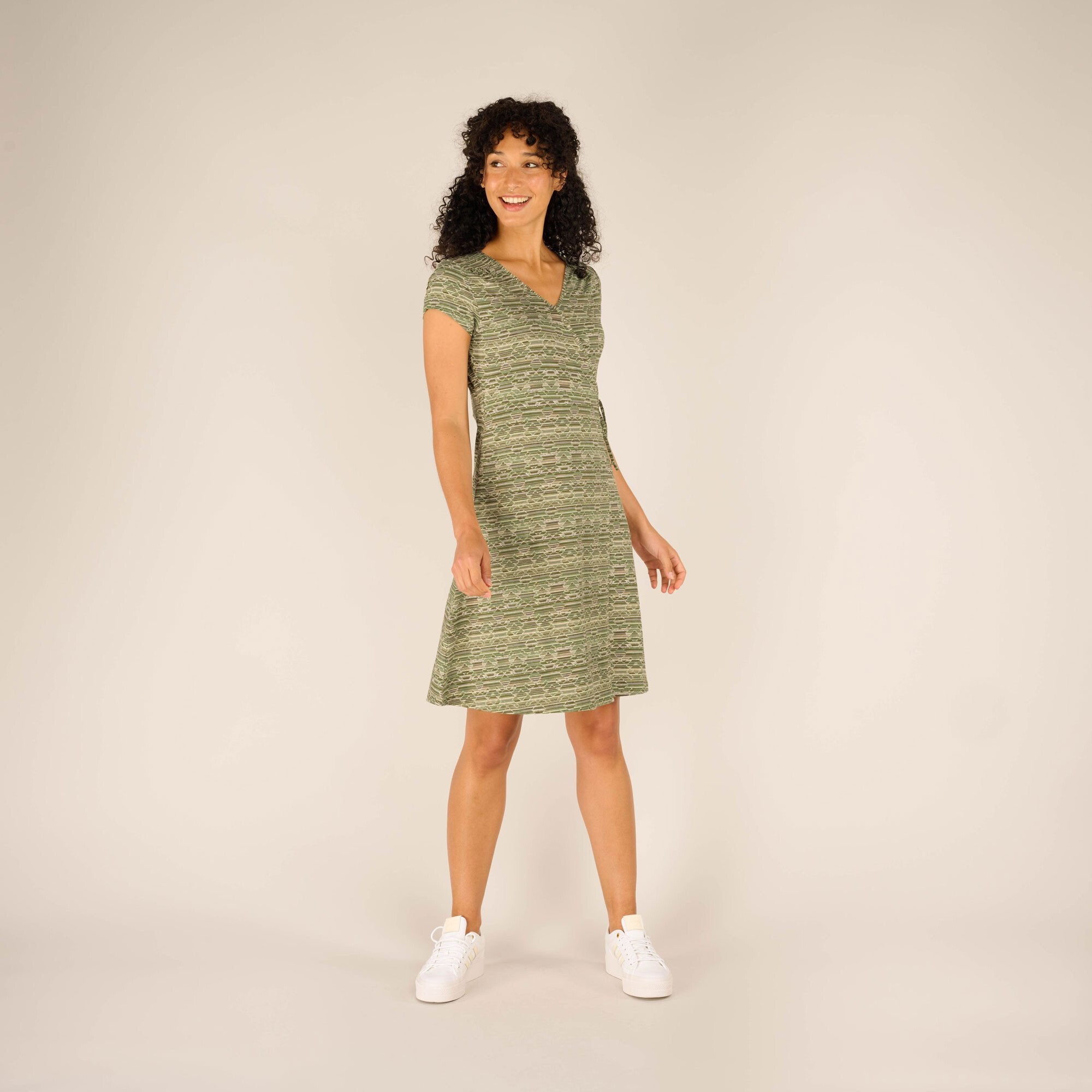 A woman with curly black hair and a radiant smile is modeling a Sherpa Adventure Gear Padma Wrap Dress in Green. The dress features a V-neckline and short sleeves, falling just above the knees. She is standing facing forward with one hand relaxed to the side. She pairs the dress with white trainers, giving a casual yet stylish look.